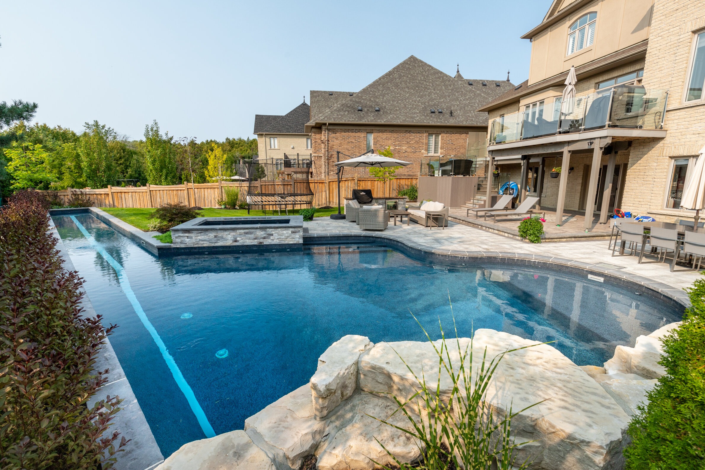 Backyard with a luxurious inground swimming pool, stone patio, outdoor furniture, lush greenery, and a two-story house with a balcony.
