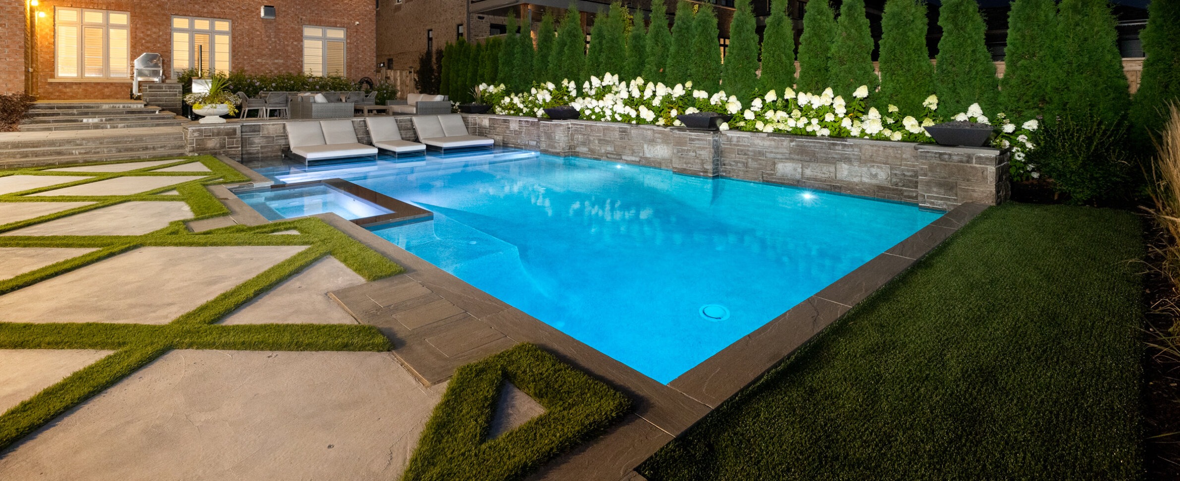 A luxurious backyard at dusk featuring an illuminated blue swimming pool, stone patio, greenery, loungers, and an adjacent brick house.