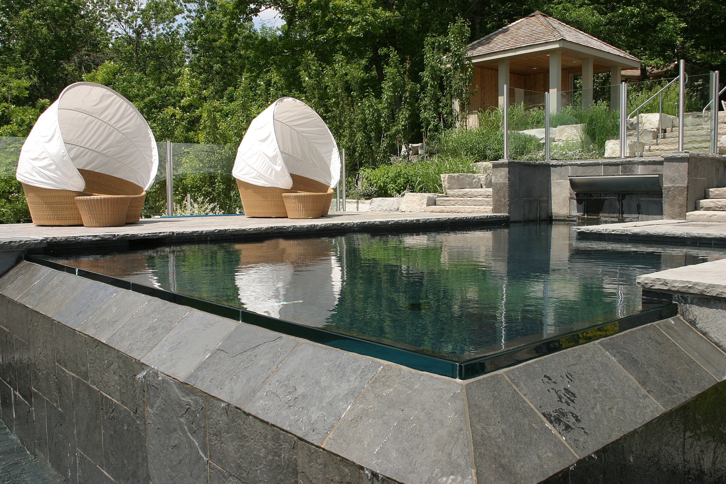 An outdoor pool with stone edges features two unique clamshell chairs and a gazebo in a lush, green landscaped backyard.