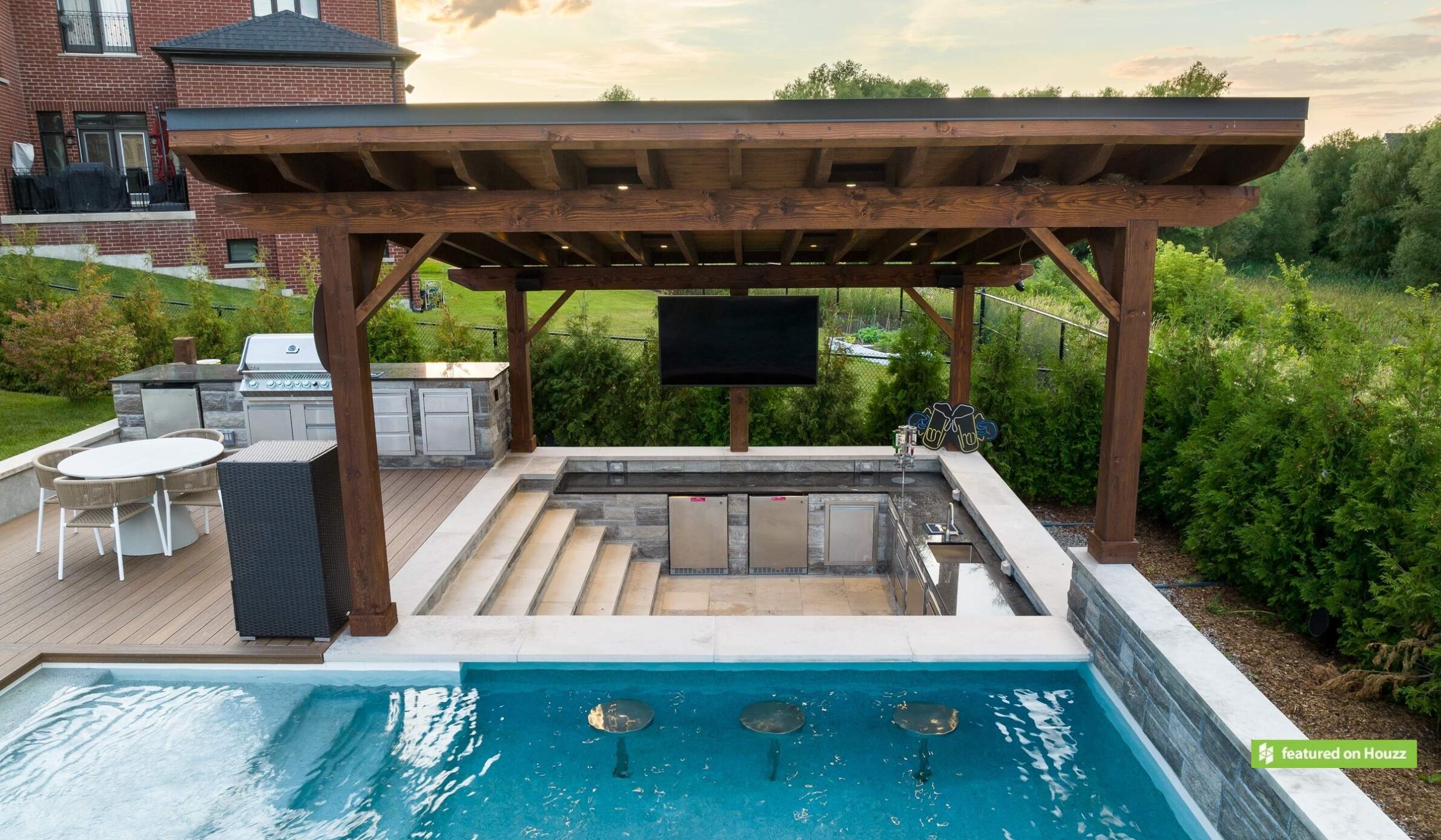 An outdoor pool with a wooden pergola, a sunken seating area, a mounted television, and a built-in grill station set amidst a landscaped background.