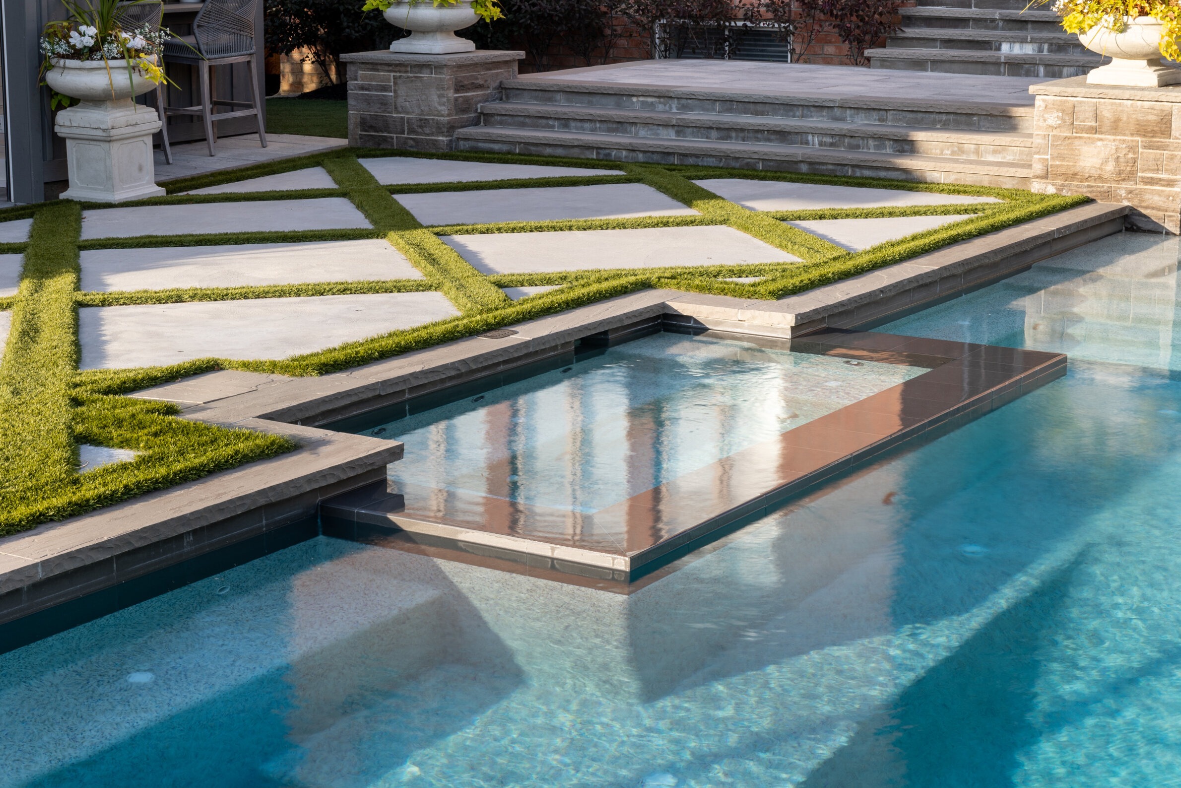 An elegant swimming pool with geometric stepping stones surrounded by manicured grass and large stone stairs, reflecting a clear blue sky.