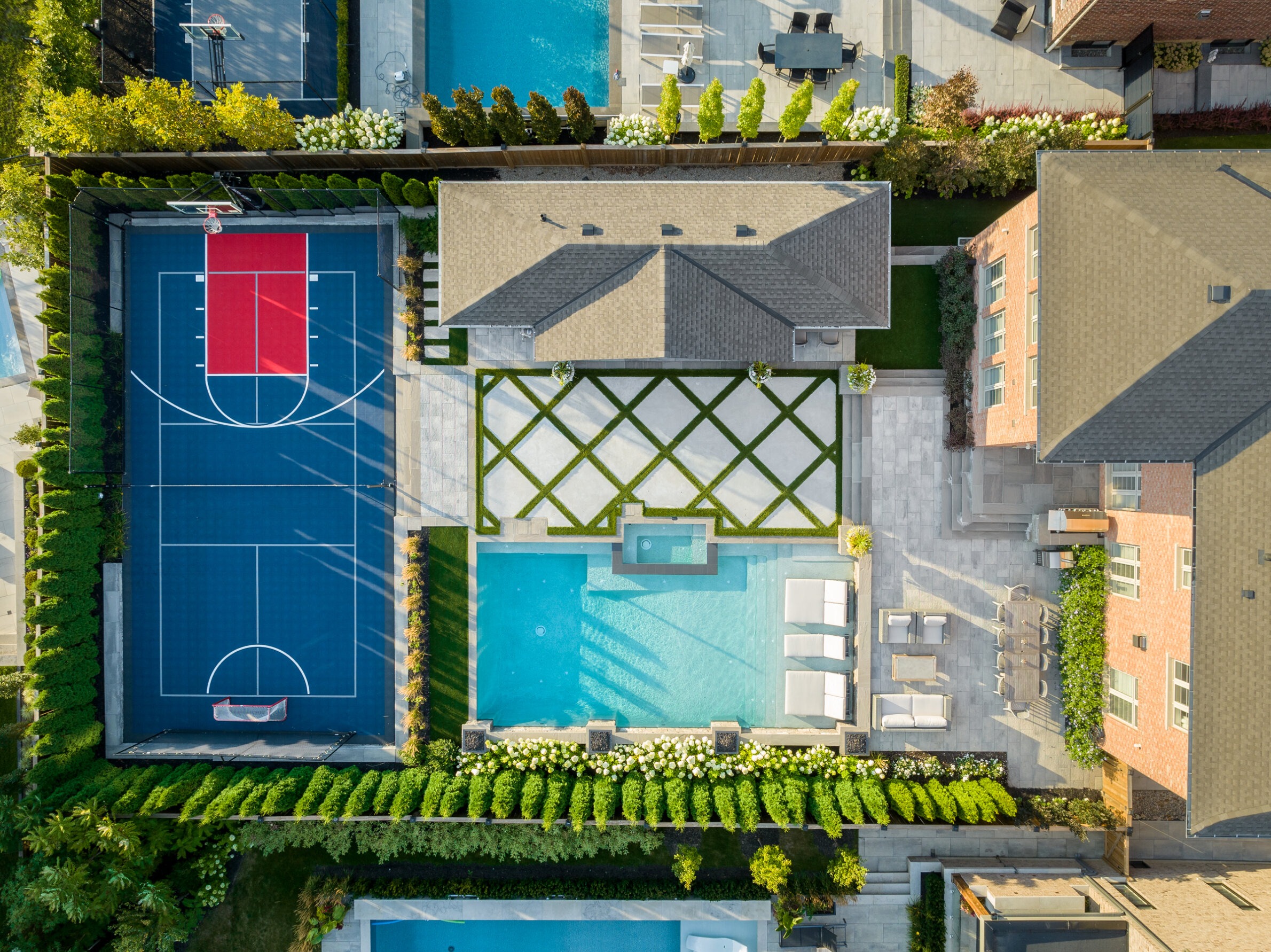 Aerial view of a luxurious backyard with a basketball court, swimming pool, landscaping, lounging chairs, and adjacent houses under clear skies.