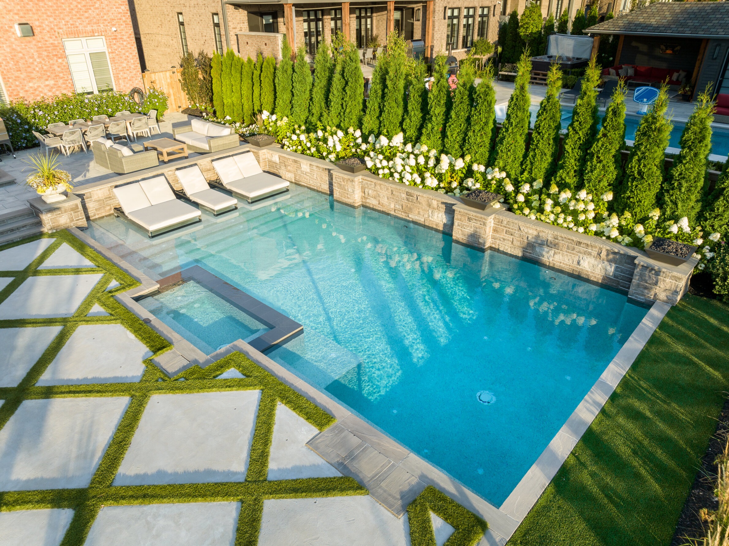 An outdoor swimming pool with clear blue water, surrounded by lounge chairs, greenery, and a well-manicured lawn with geometric patterns.