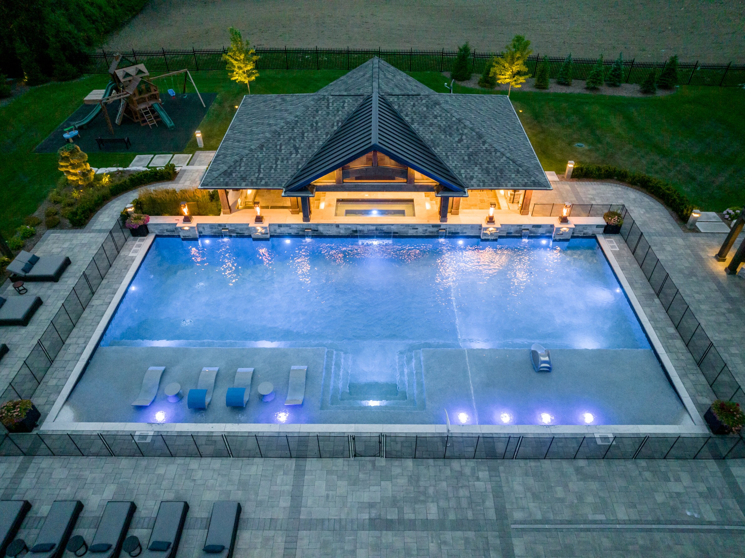 Aerial view at dusk of a luxurious backyard with a large swimming pool, lounge chairs, landscaped garden, and a children