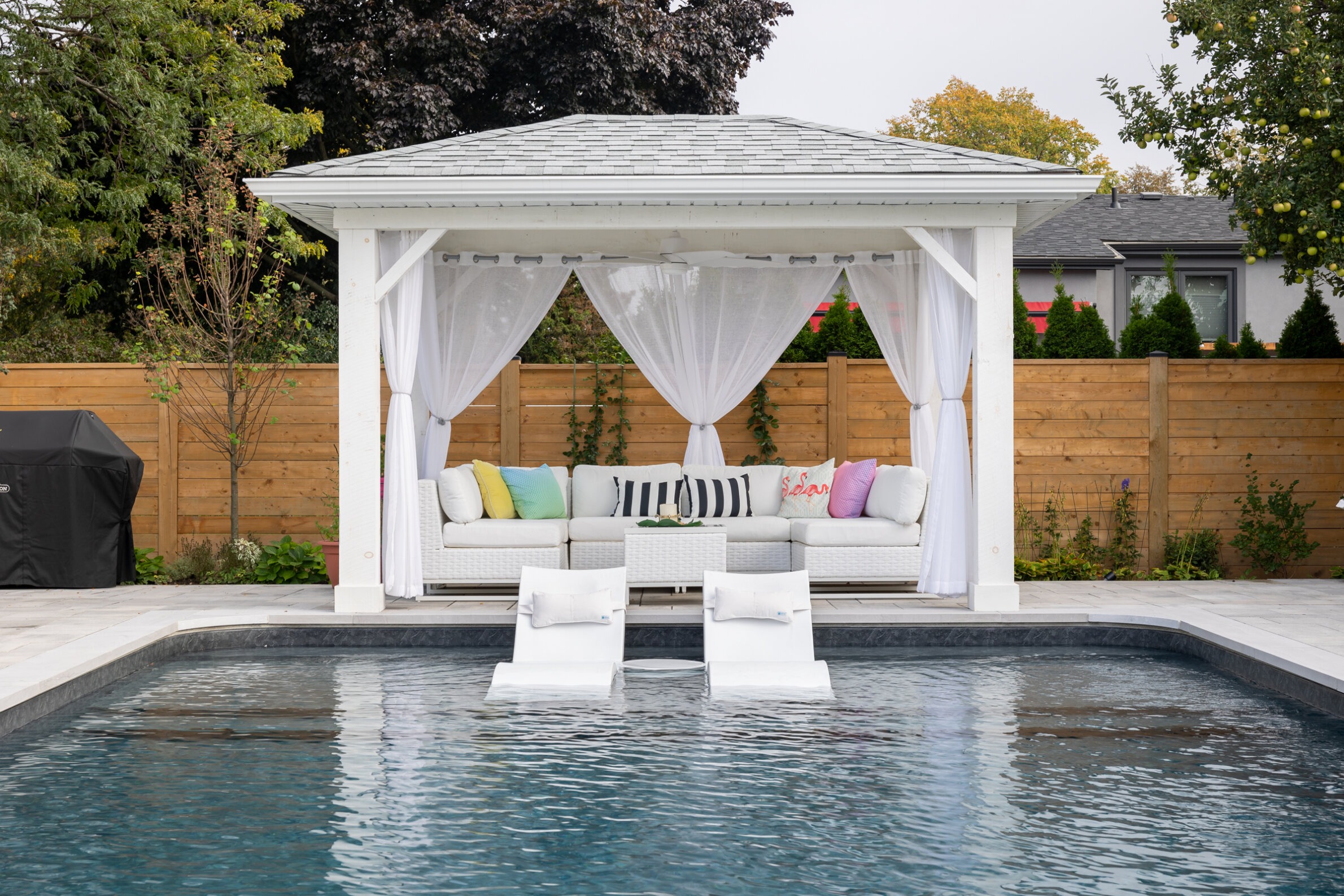 This image showcases a serene backyard with an elegant white poolside gazebo adorned with sheer curtains and colorful cushions on a sectional sofa.