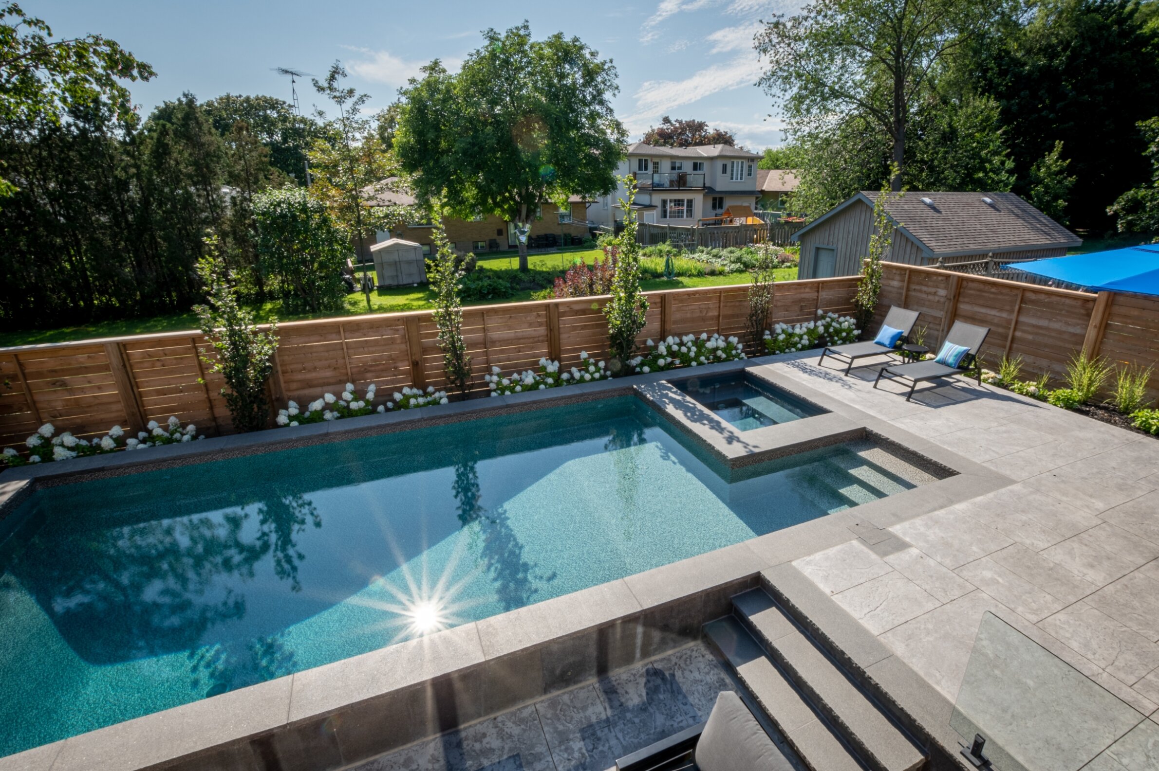 A sunny backyard with a rectangular swimming pool, lounging chairs, wooden fence, lush greenery, and a blue pool cover partially extended.