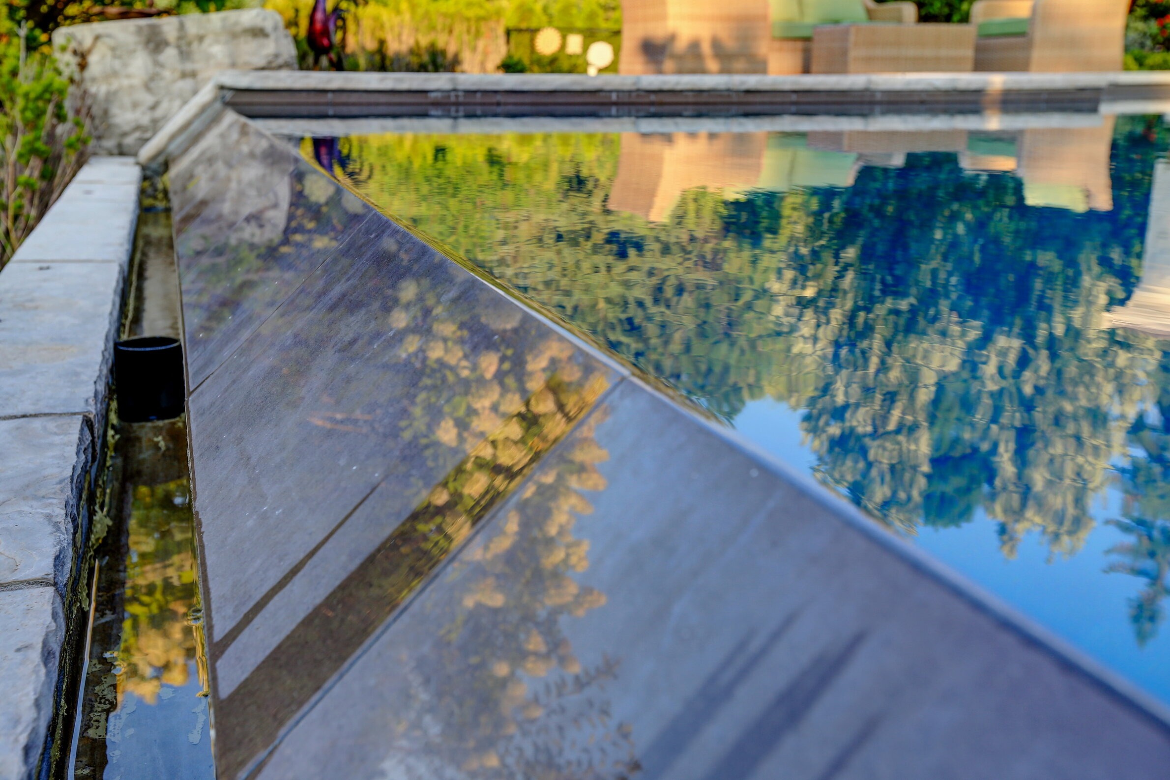 Close-up of an infinity pool's edge with reflective water mirroring trees and clear sky, adjacent to outdoor furniture on a sunny day.