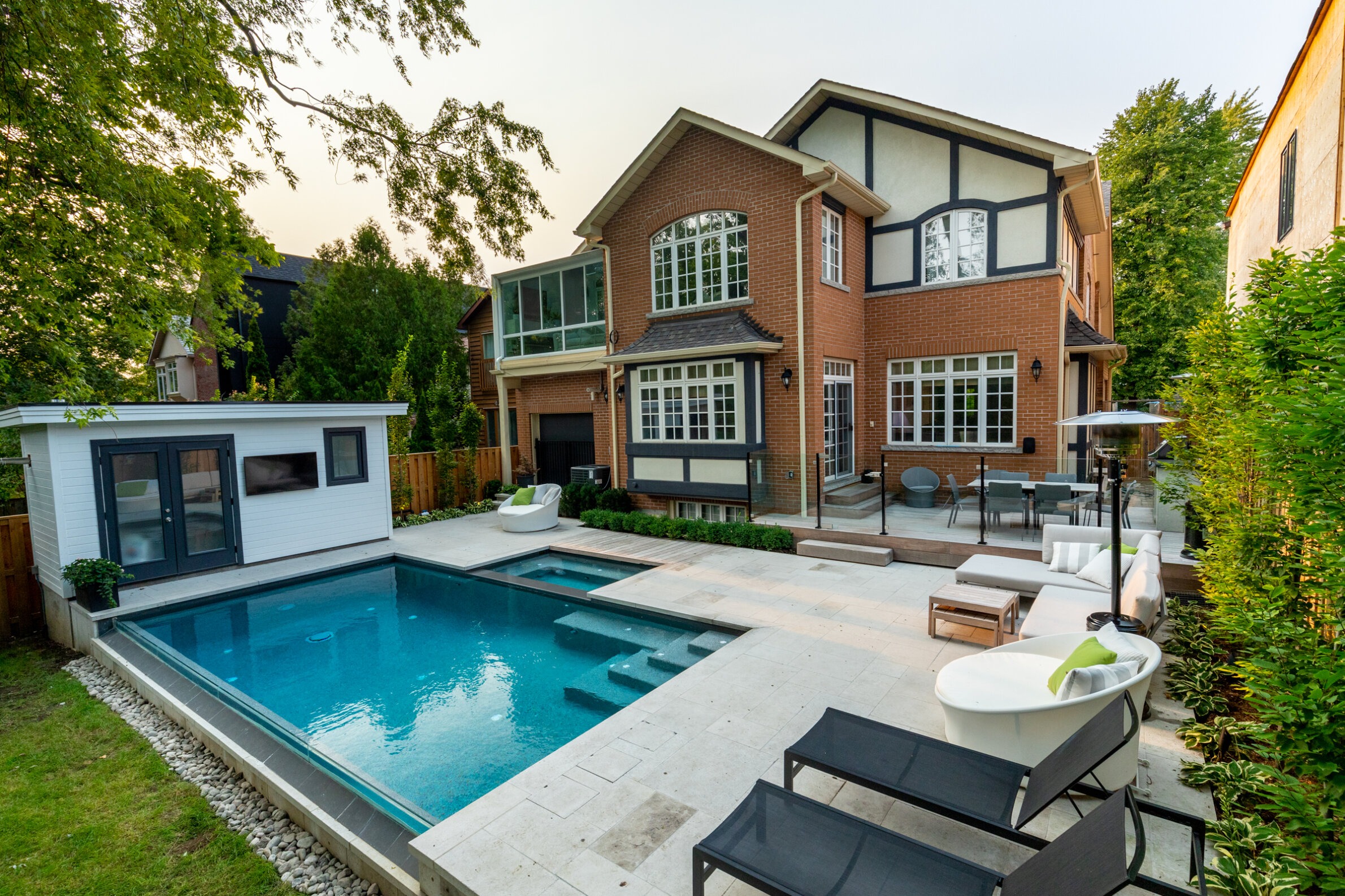 This image shows a luxurious backyard with a swimming pool, a small pool house, modern patio furniture, and a two-story brick house with large windows.