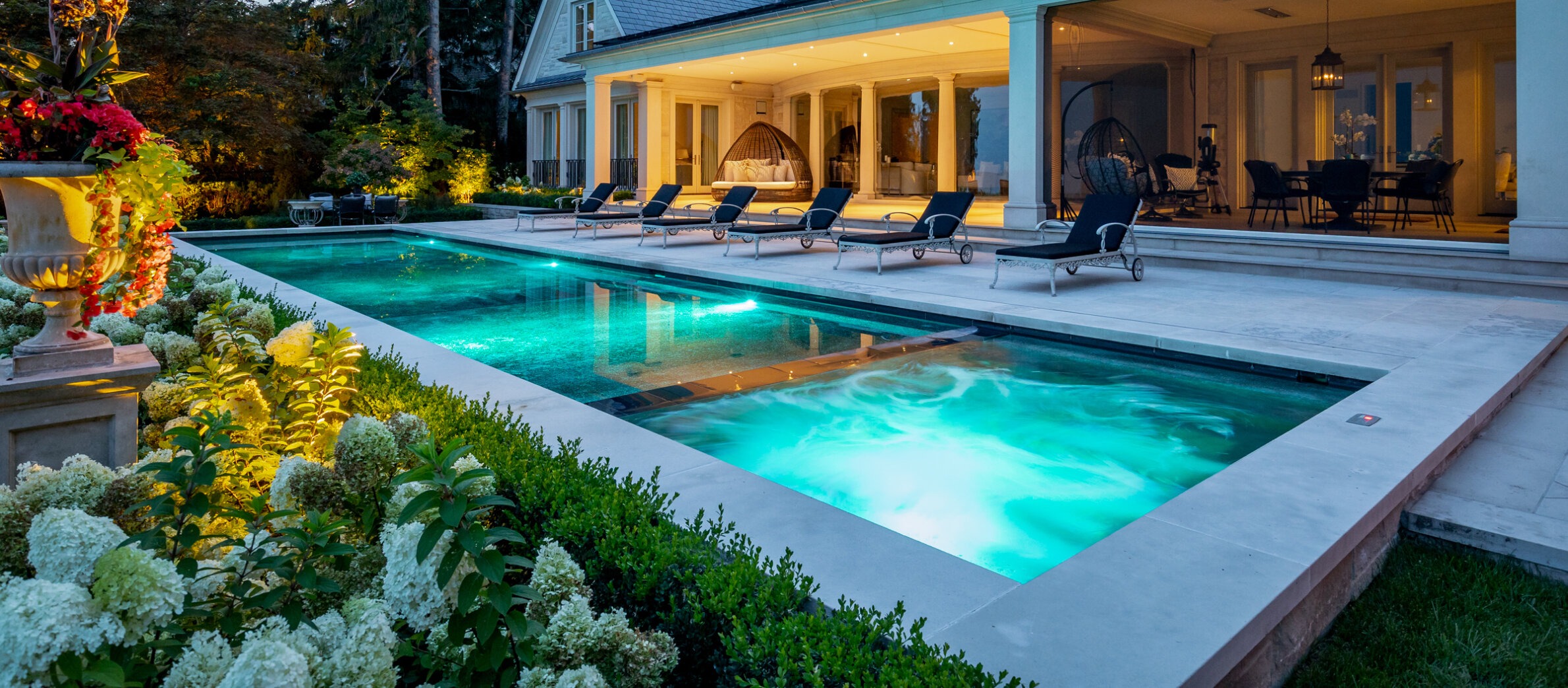 An illuminated swimming pool at dusk, surrounded by lush vegetation, fronting a luxurious house with outdoor seating and a cozy interior visible.