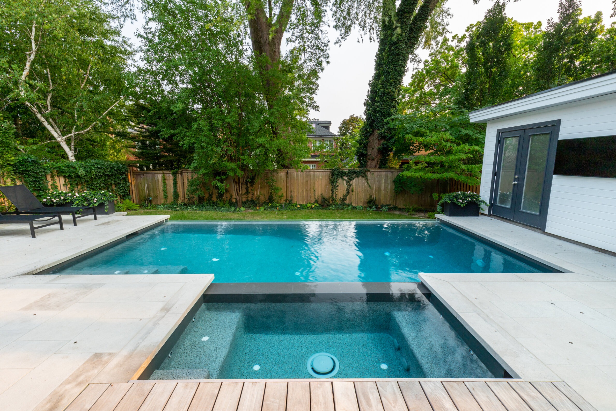 An outdoor swimming pool with an attached hot tub, surrounded by a wooden deck, lounge chair, lush trees, and a fence. A modern house is visible.