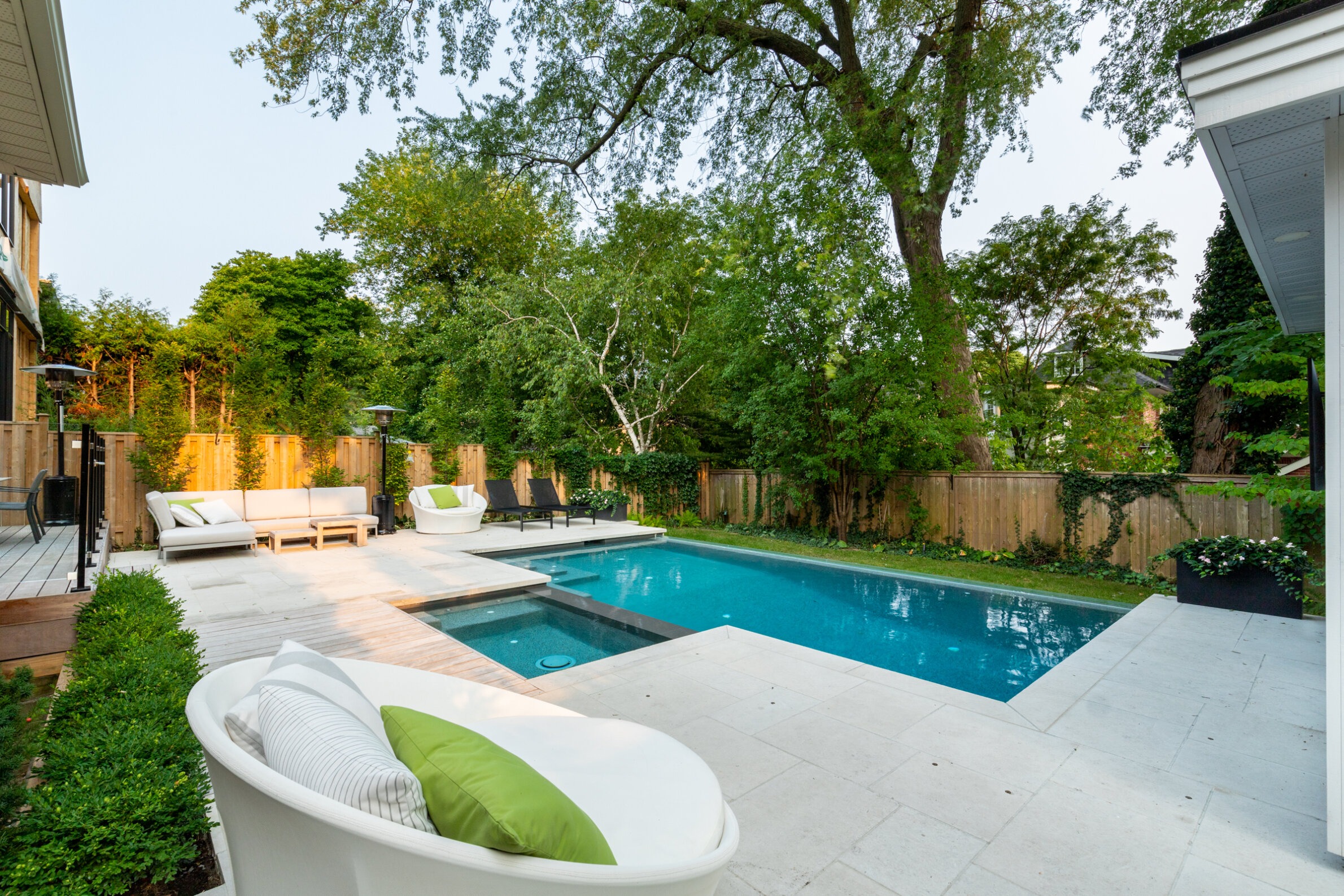 This image shows a serene backyard with a swimming pool, patio lounging area, manicured hedges, tall trees, and a wooden privacy fence.