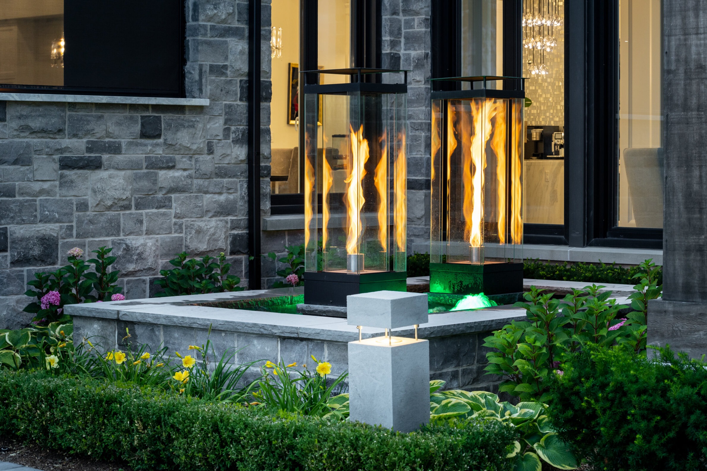 This image shows an elegant outdoor space with two large flame lanterns, greenery, and a modern house facade with reflective windows in the background.