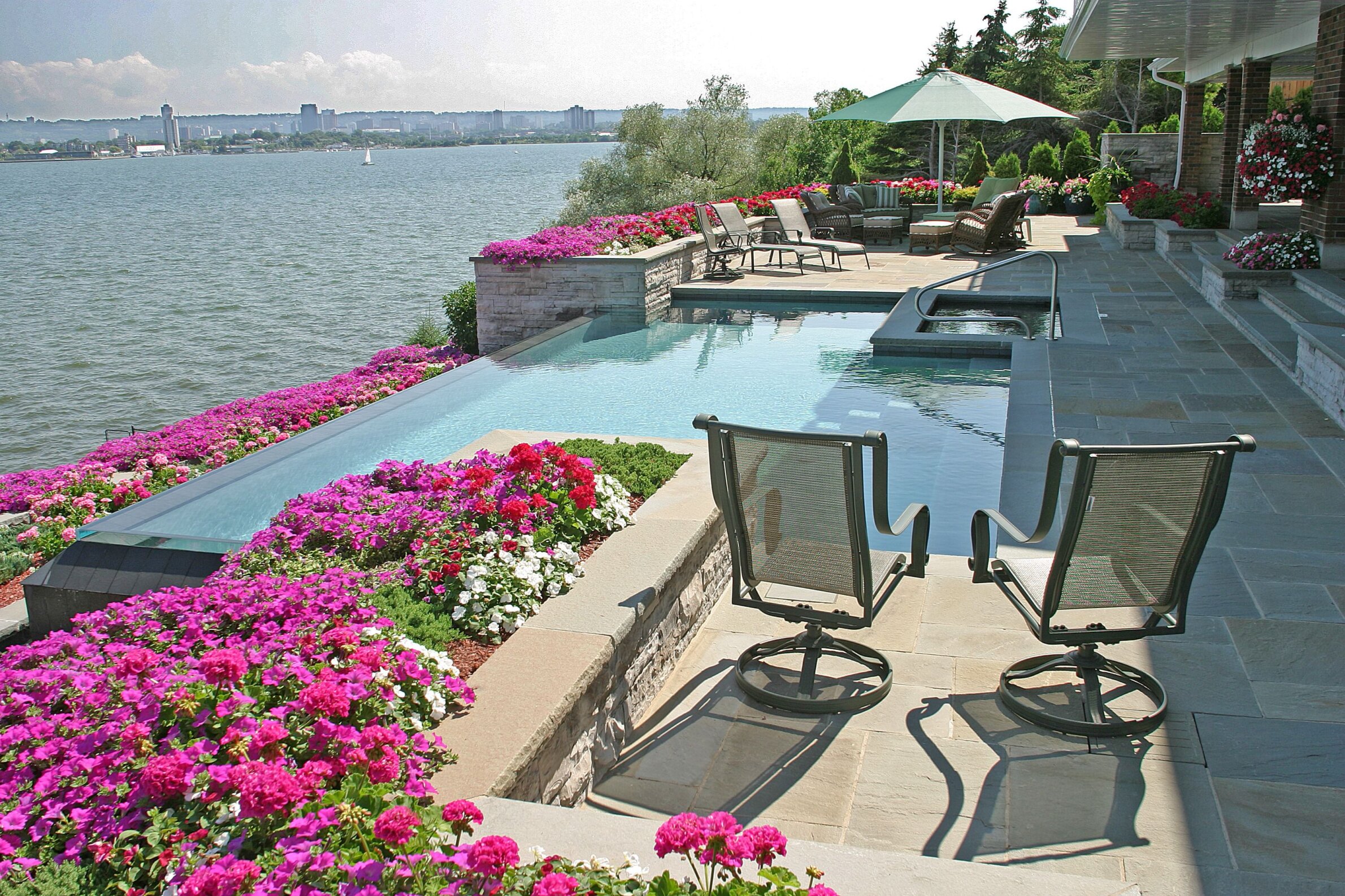 A luxurious outdoor pool area with abundant flowers, patio furniture, and a scenic waterfront view under a sunny sky.