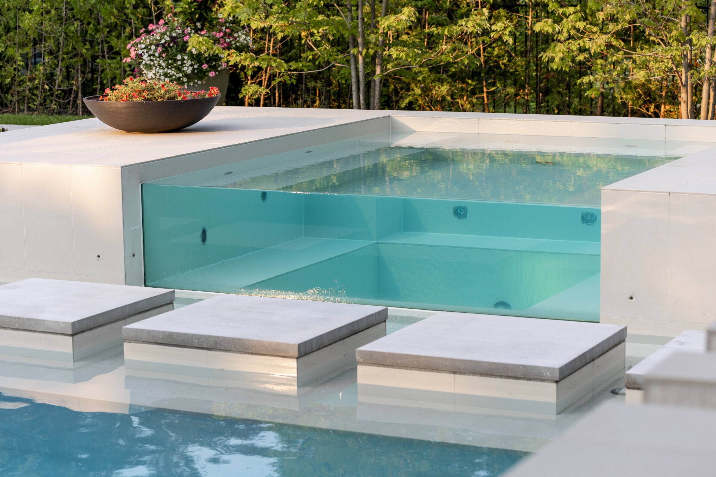 A modern outdoor swimming pool with clear blue water, surrounded by white decking and stepping stones, adjacent to a lush green backdrop.