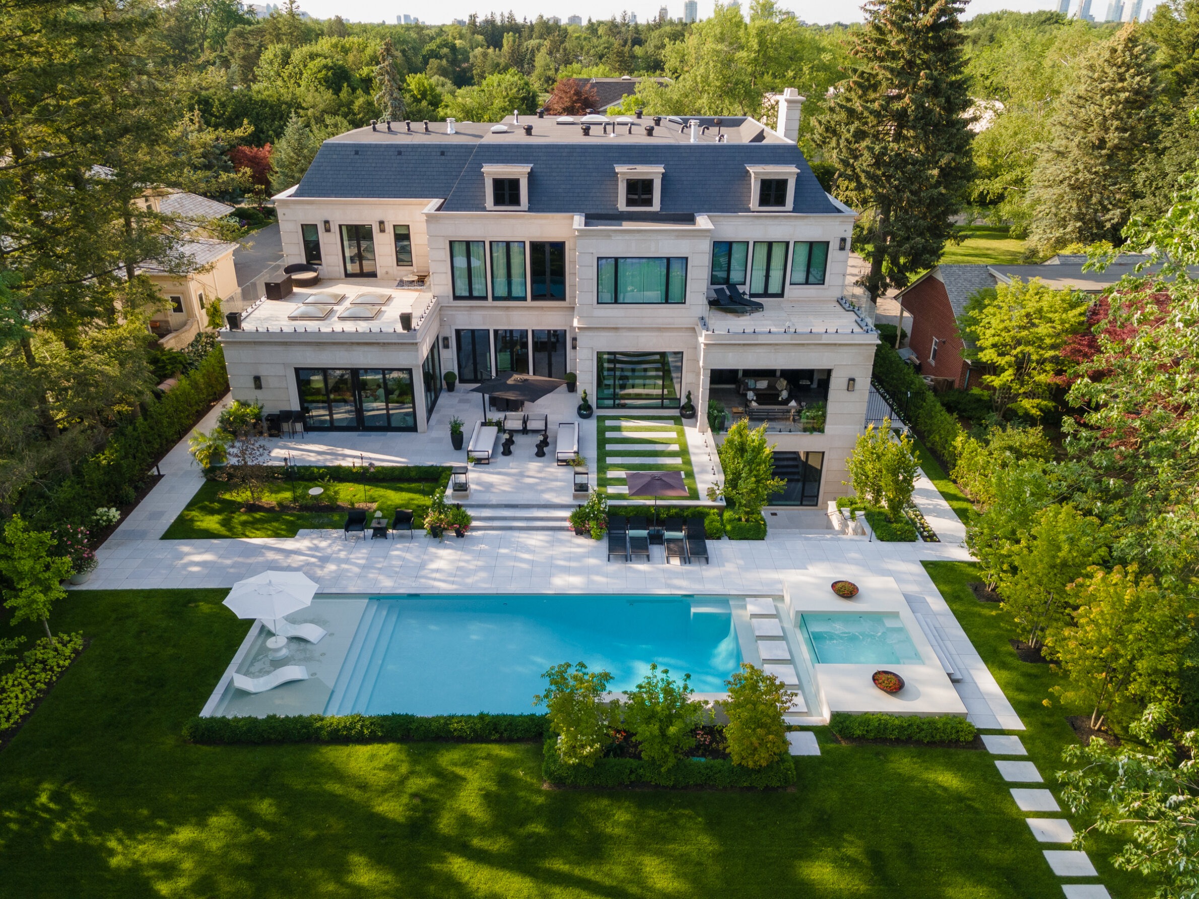 Aerial view of a large, luxurious house with a neatly landscaped backyard, featuring a swimming pool, outdoor seating, and surrounding greenery.