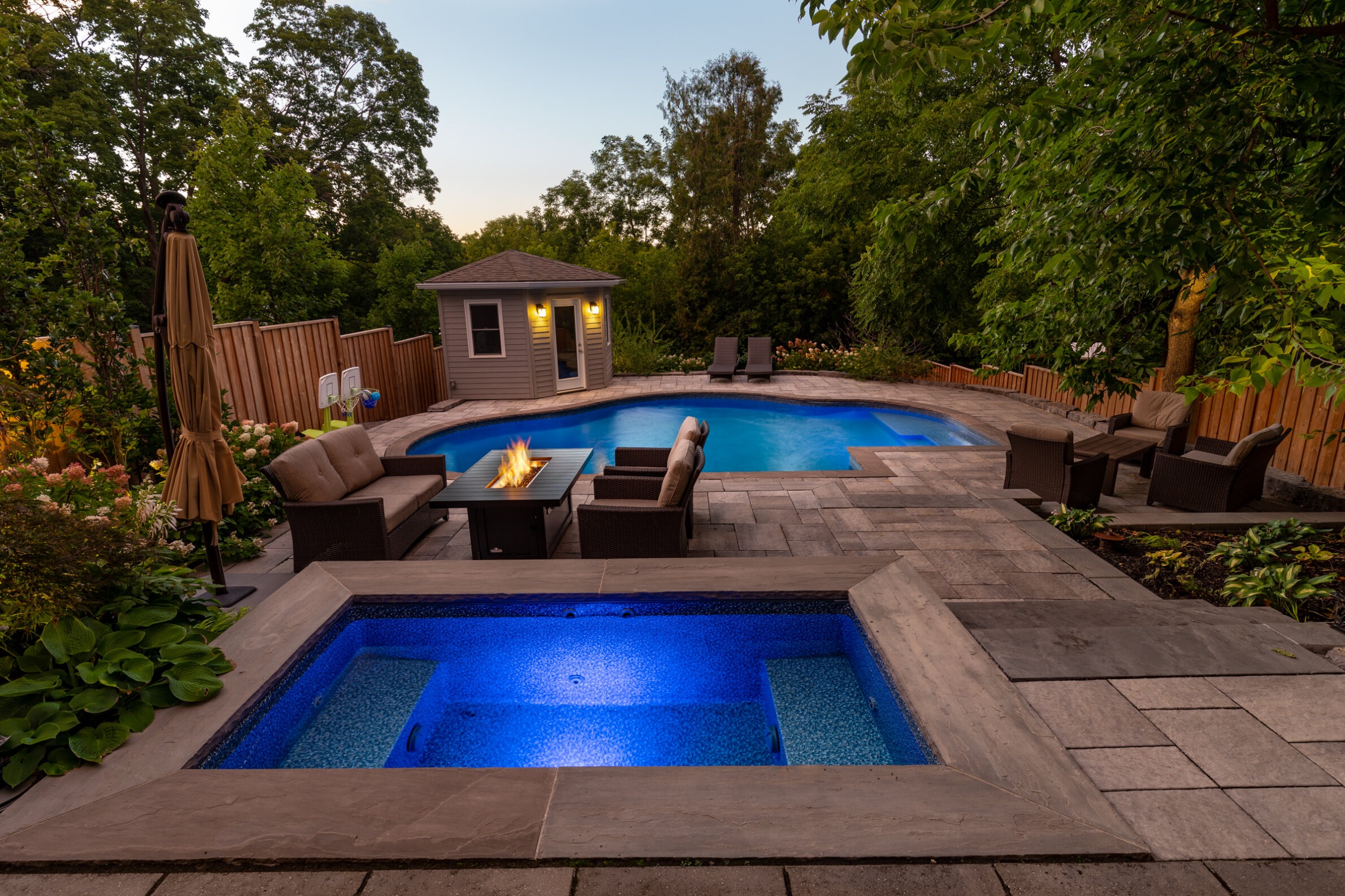 A tranquil backyard with a swimming pool, spa, outdoor furniture, gas firepit, neatly landscaped plants, wooden fence, and a small pool house at dusk.