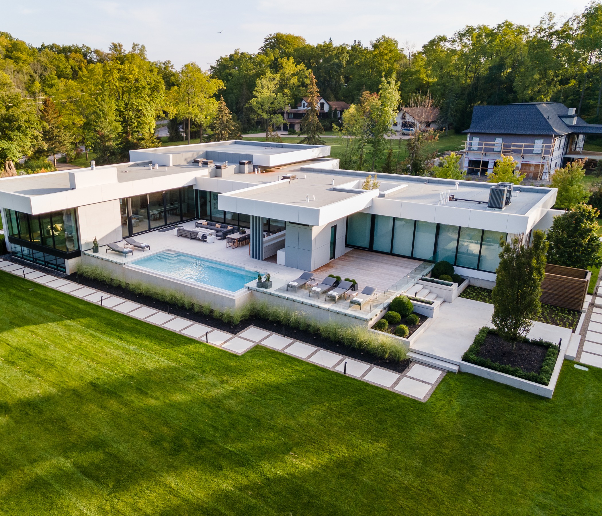 An aerial view showcases a luxurious modern home with flat roofs, expansive glass walls, an outdoor swimming pool, and well-manicured lawn and hedges.