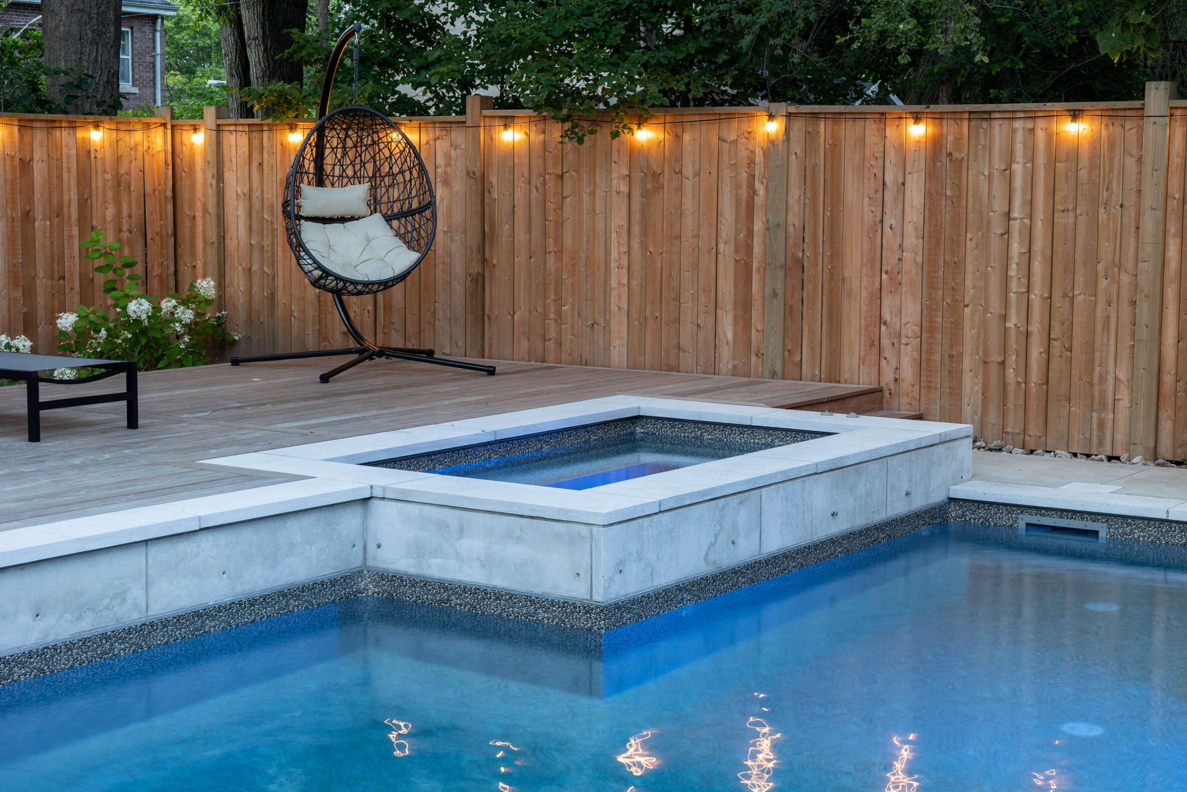 An outdoor area featuring a swimming pool with blue water, surrounded by a deck, a hanging chair, and a wooden fence with string lights.