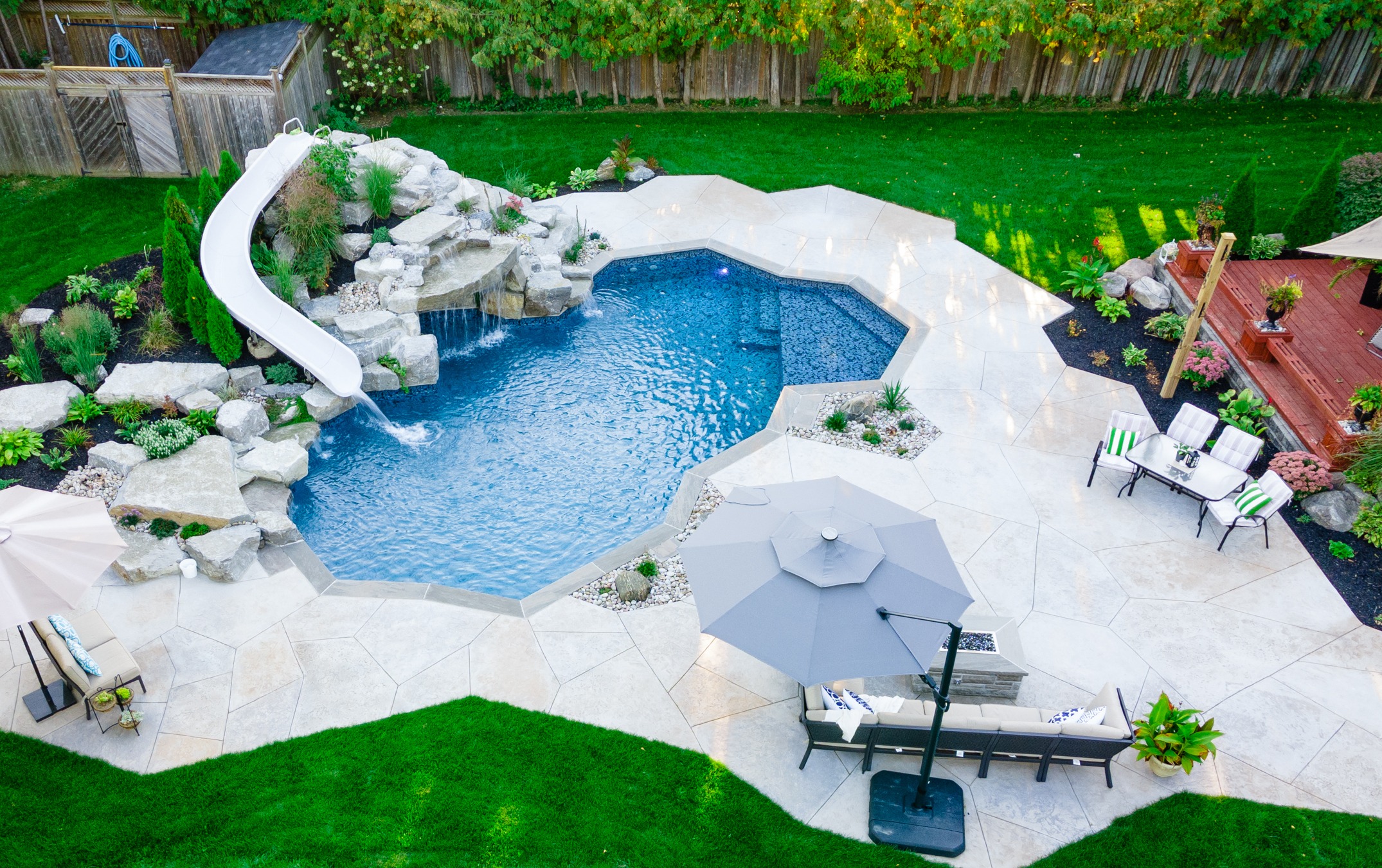 Aerial view of a landscaped backyard with a blue swimming pool, white waterslide, surrounding stone features, patio area, green lawn, and wooden deck.