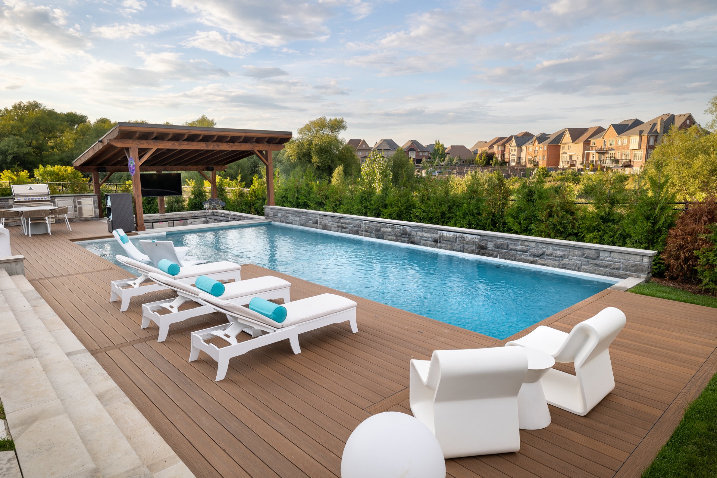 This is an outdoor residential pool area featuring lounge chairs, a pergola, and a built-in grill, with a backdrop of suburban houses and trees.