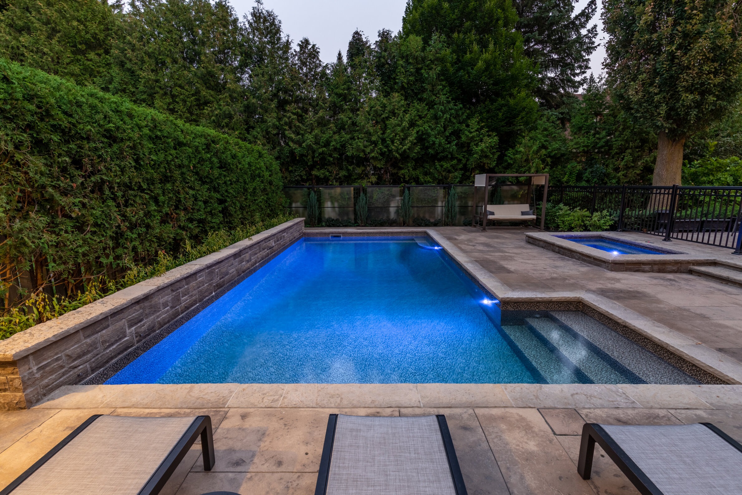 An outdoor swimming pool glows blue at dusk, surrounded by greenery and loungers, with a hot tub, steps, and a fence visible.