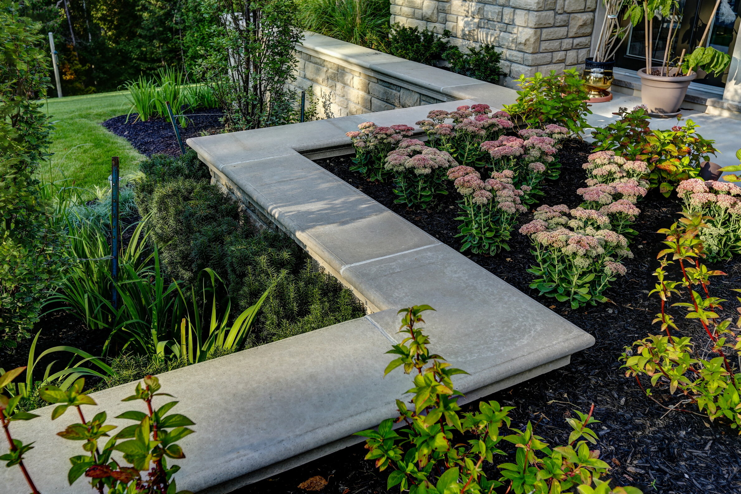 A landscaped garden featuring stone pathways, blooming pink flowers, lush greenery, and a neatly mulched bed with a stone wall and potted plant.