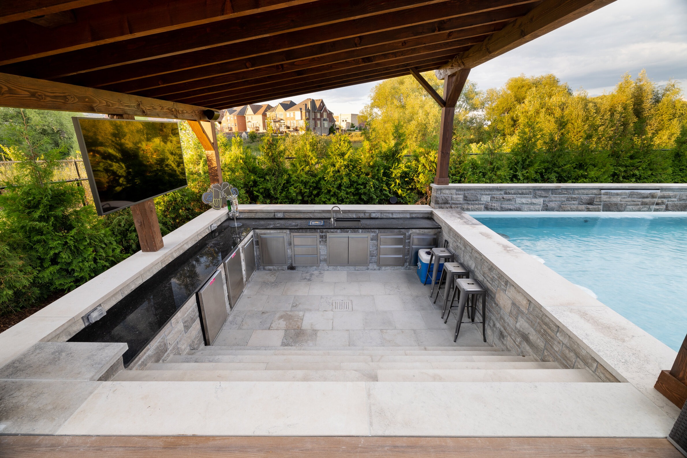An outdoor kitchen with stainless steel cabinets adjacent to a clear, blue swimming pool. Wooden steps lead down from a covered patio area.