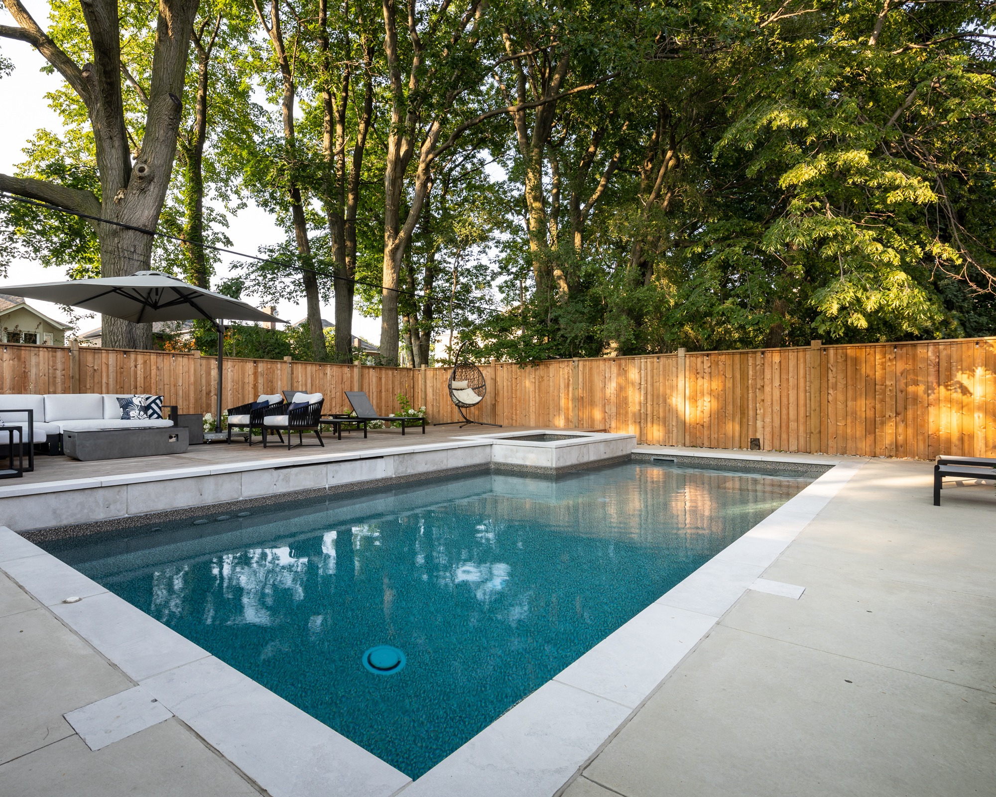 This image shows a tranquil backyard with a swimming pool, modern patio furniture, trees, and a wooden fence illuminated by warm sunlight.