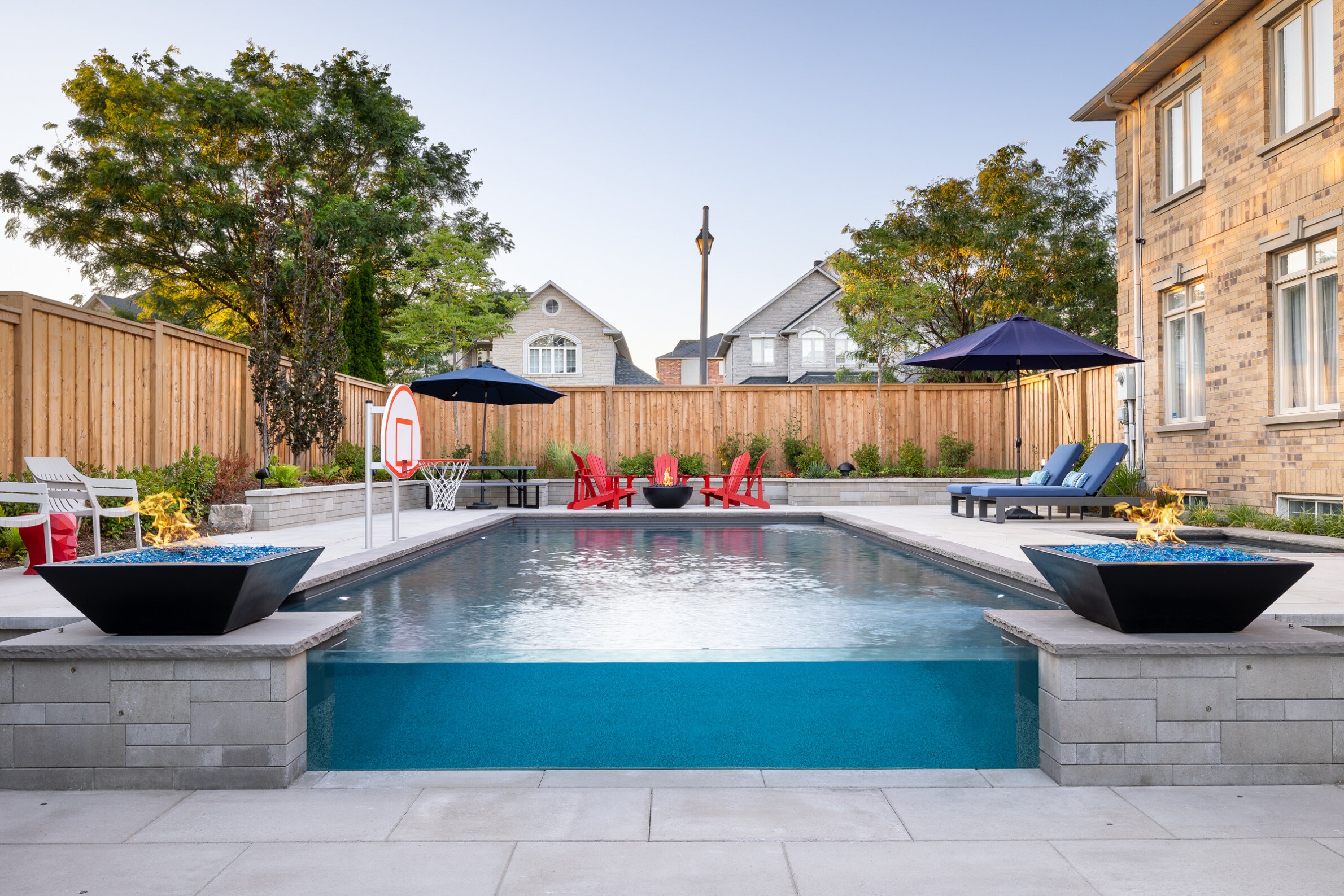 A backyard setting with an inground pool, surrounded by patio chairs, umbrellas, a basketball hoop, and a fire feature, with a wooden fence backdrop.