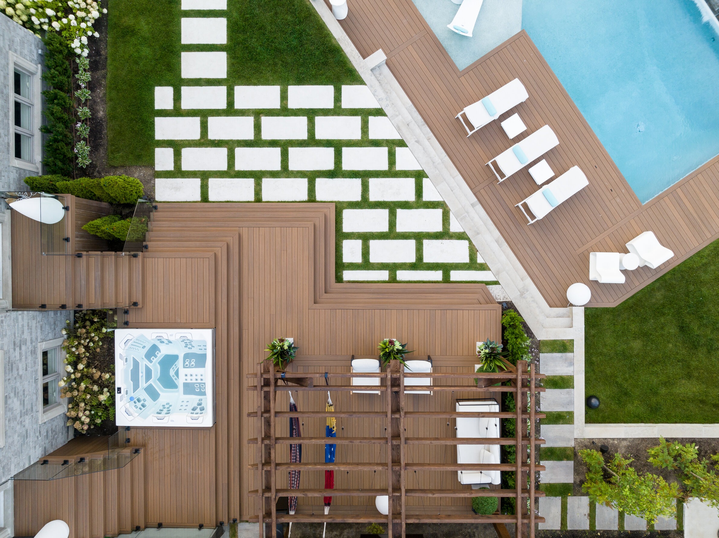 Aerial view of a backyard with a pool, sun loungers, hot tub, patterned grass squares, wood decking, and neatly landscaped plants.