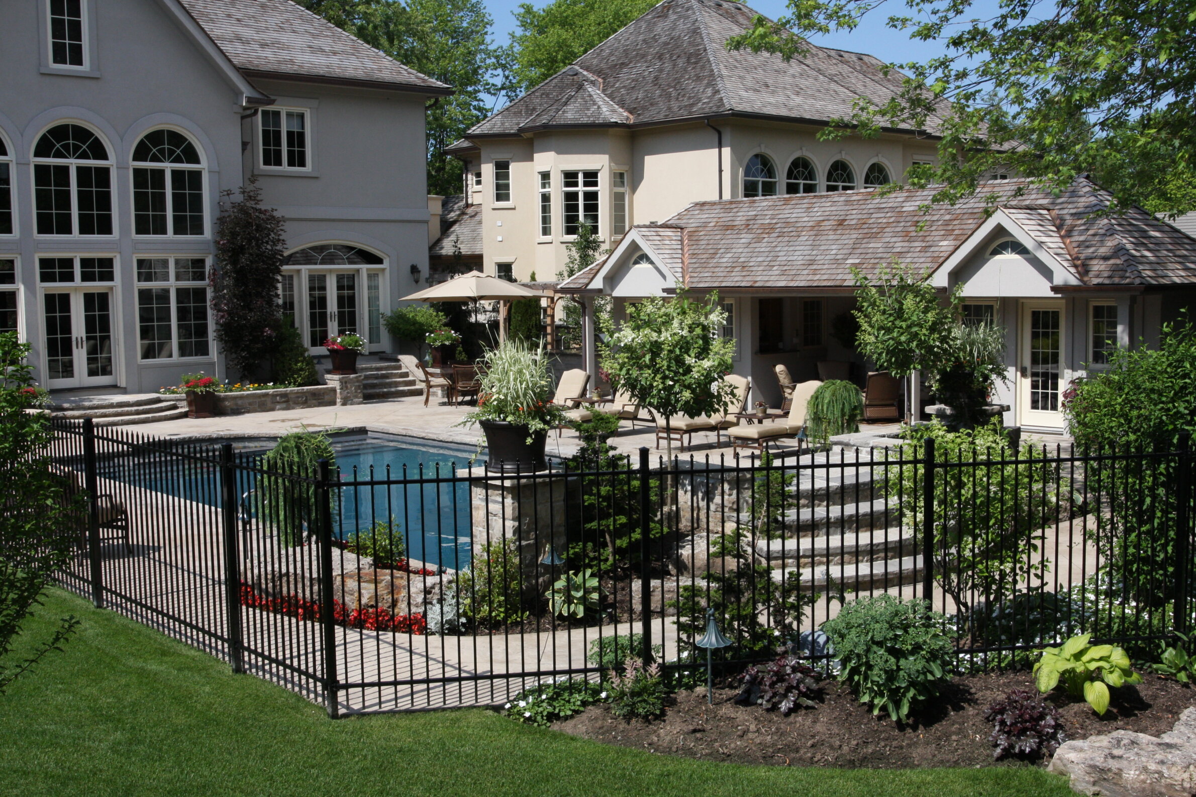 An upscale backyard with a swimming pool, neatly manicured lawn, patio furniture, and a grand house featuring large windows and multiple rooflines.