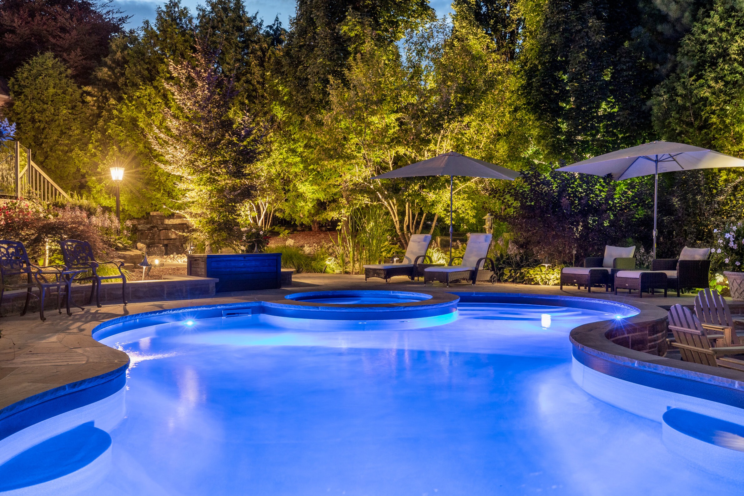 An evening view of a serene backyard with an illuminated swimming pool, hot tub, lounge chairs, patio furniture, and well-landscaped garden with trees.