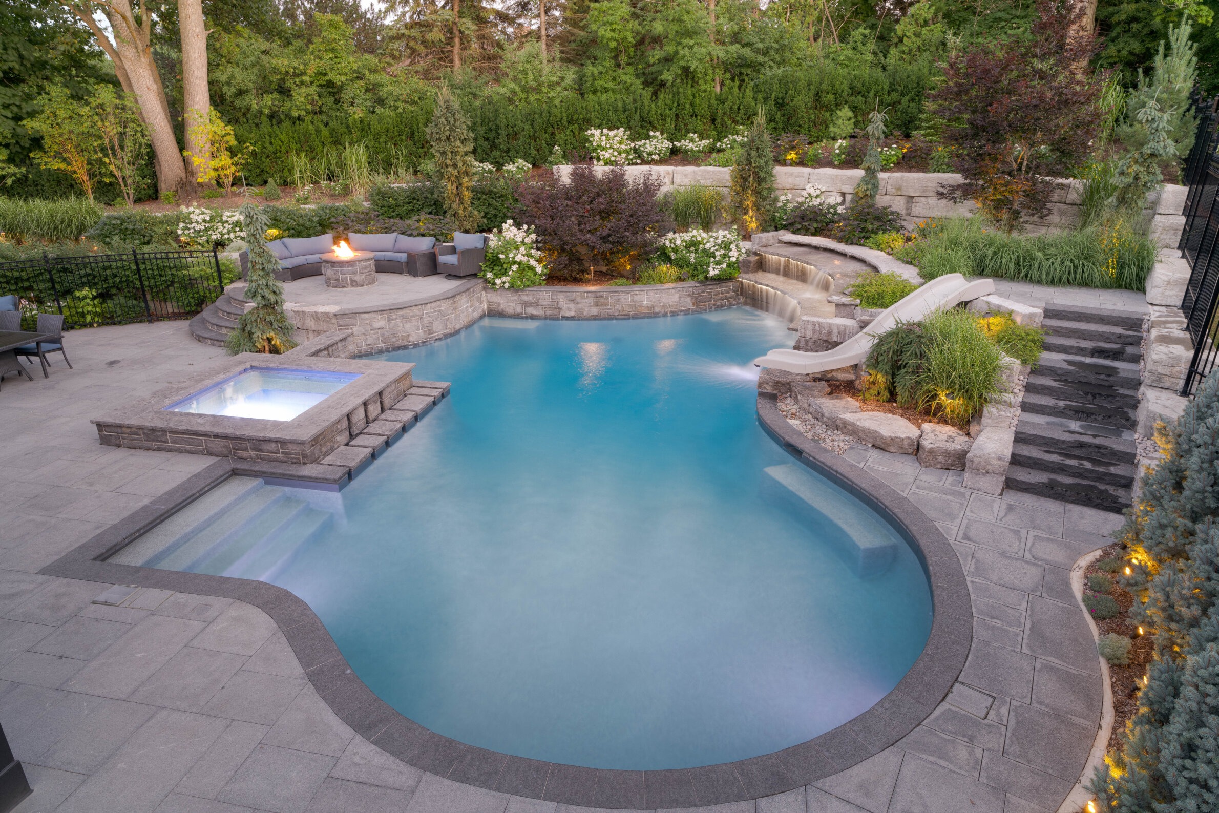 This image shows a luxurious backyard with a curved swimming pool, hot tub, stone patio, fire pit, and a waterslide amid landscaped greenery at dusk.