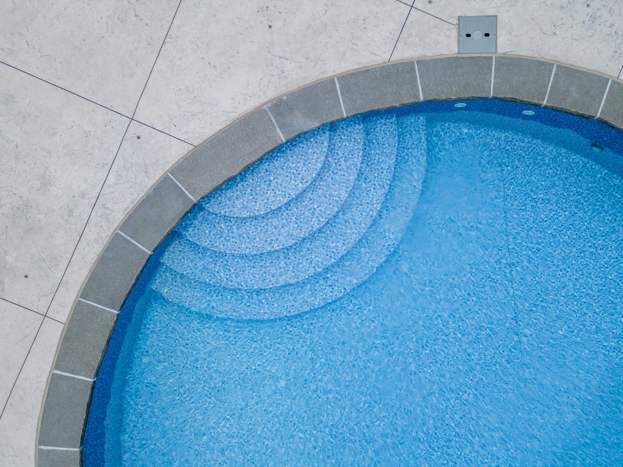 This is an aerial view of a corner of a circular pool with steps leading into the water, surrounded by a tiled surface, with a pool light visible.