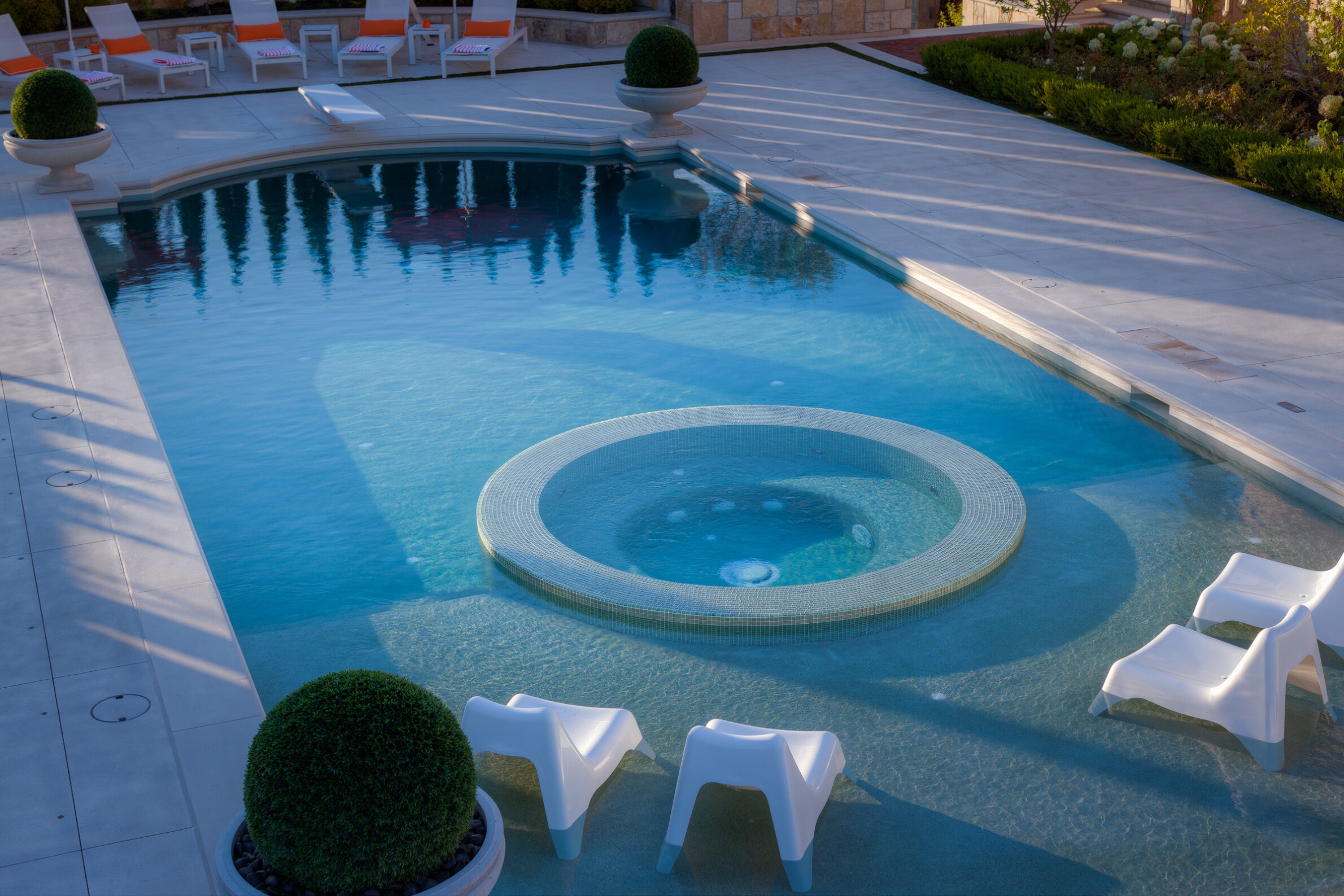 A tranquil swimming pool with a built-in jacuzzi, surrounded by lounge chairs and manicured shrubs, reflects a clear blue sky.