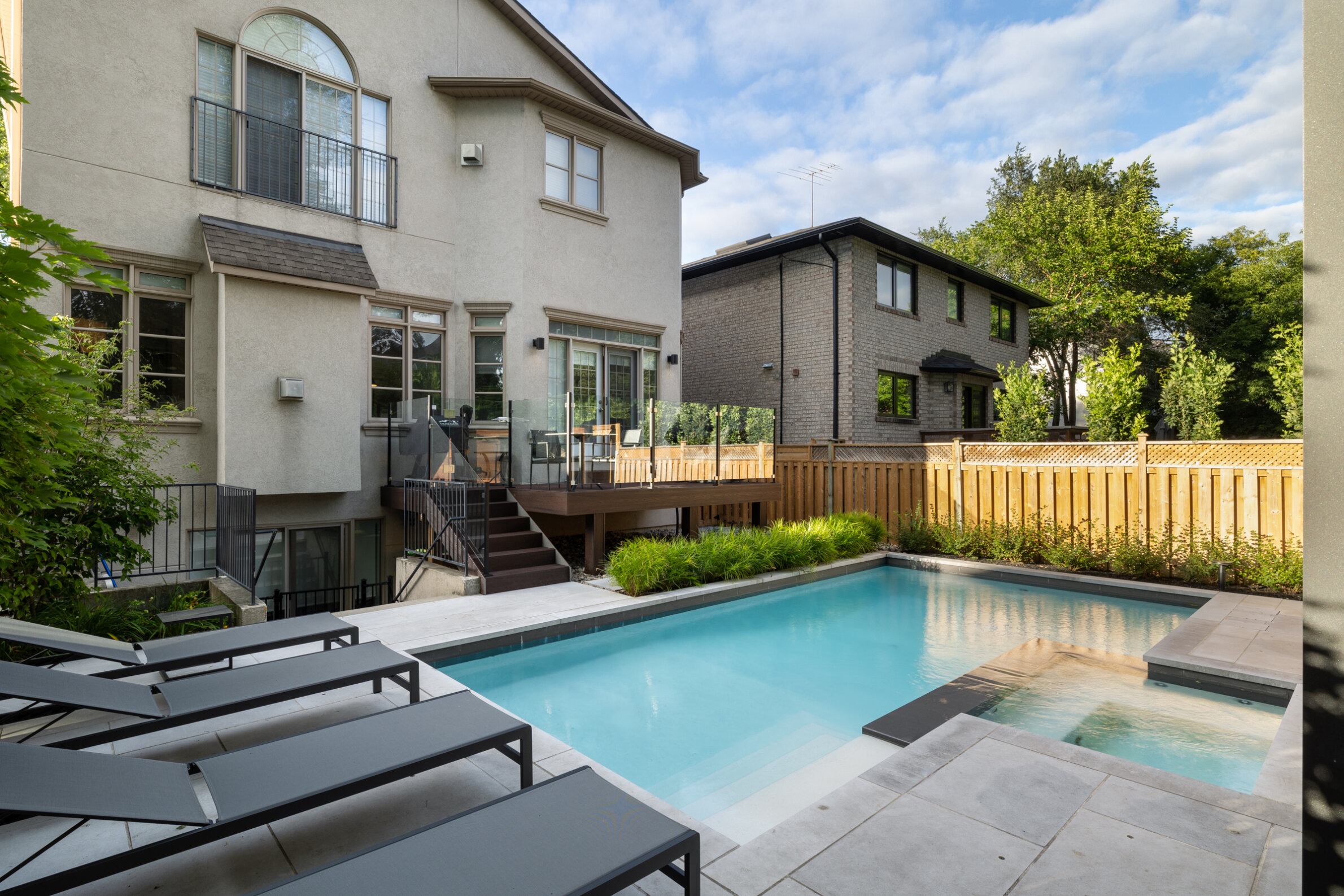 A residential backyard with a clear pool, patio area, outdoor furniture, wooden fence, and two-story houses surrounded by greenery under a partly cloudy sky.