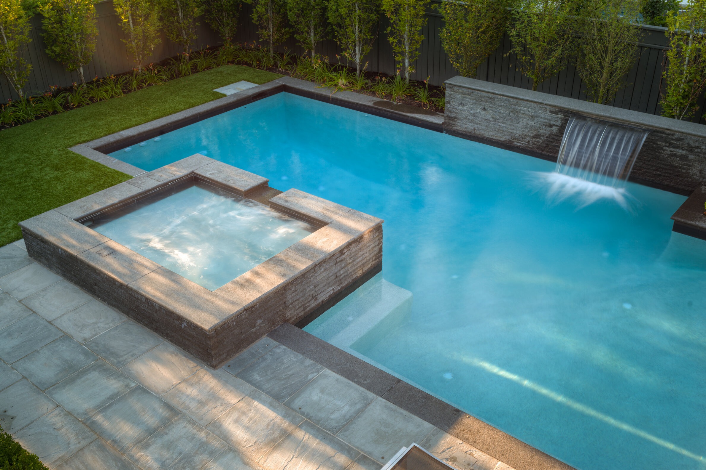 A serene backyard featuring a large swimming pool with a waterfall and an attached hot tub, surrounded by a lush lawn and privacy fencing.
