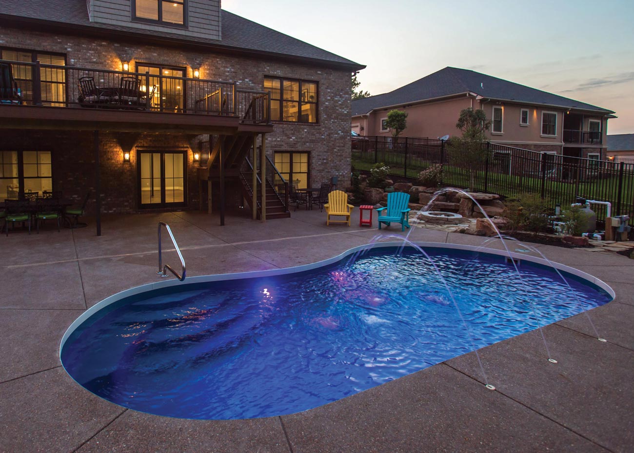 A residential backyard at dusk featuring a kidney-shaped pool, a two-story house with lit windows, and colorful patio chairs on a paved surface.