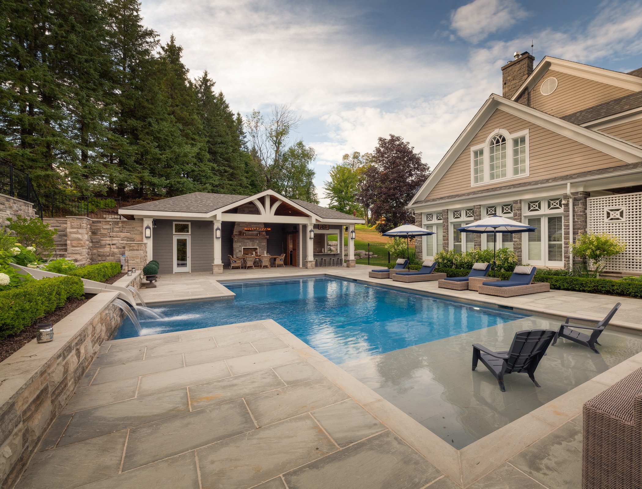 This image shows a luxurious backyard with a swimming pool, sun loungers, pool house, and a well-maintained garden against a backdrop of trees.