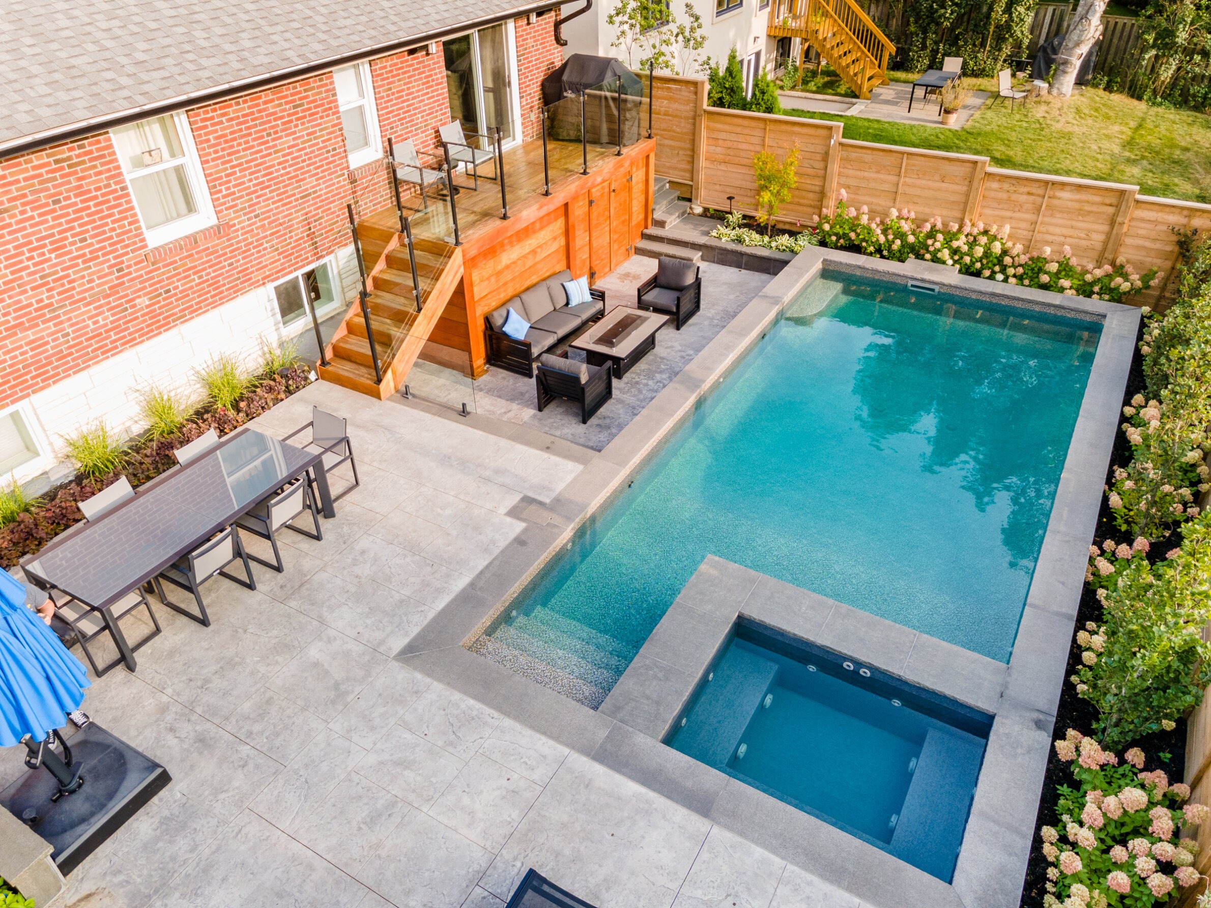 Aerial view of a backyard with a rectangular swimming pool, hot tub, patio area with furniture, lawn, and a person standing by the table.
