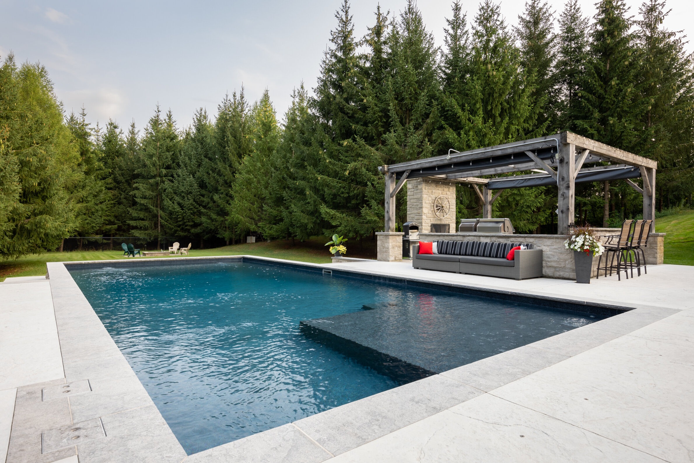 An outdoor swimming pool with lounge area and pergola set against a backdrop of tall green trees under a cloudy sky. Modern, tranquil, and luxurious setting.