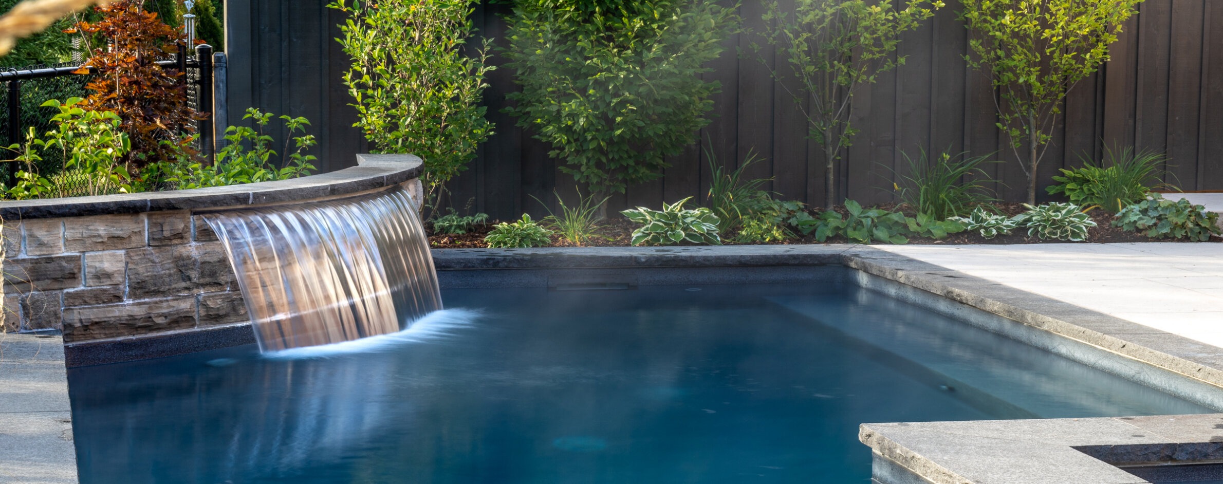 An outdoor swimming pool with a smooth waterfall edge, surrounded by lush greenery and a dark fence, depicting a serene backyard setting.