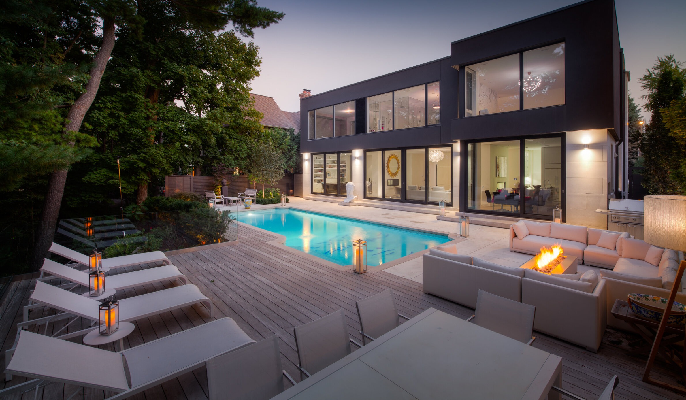 Modern house with large windows at dusk, featuring a swimming pool, outdoor seating with a fire pit, and lush surrounding greenery.