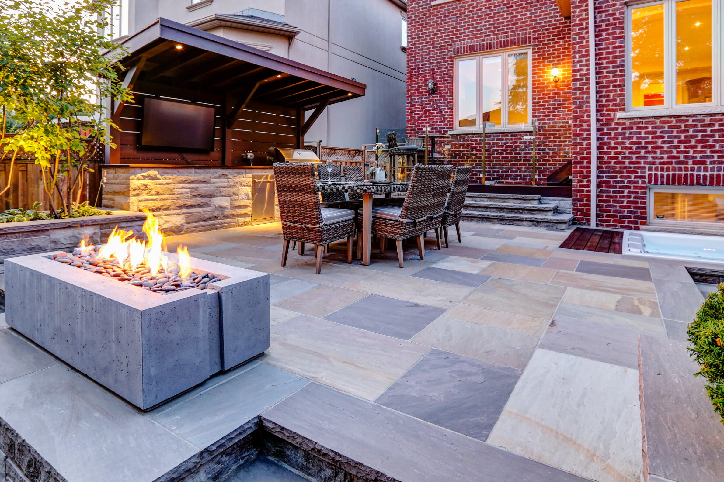 This image shows an outdoor patio with a fire pit, dining furniture, a pergola with a mounted TV, and a hot tub beside a brick house.
