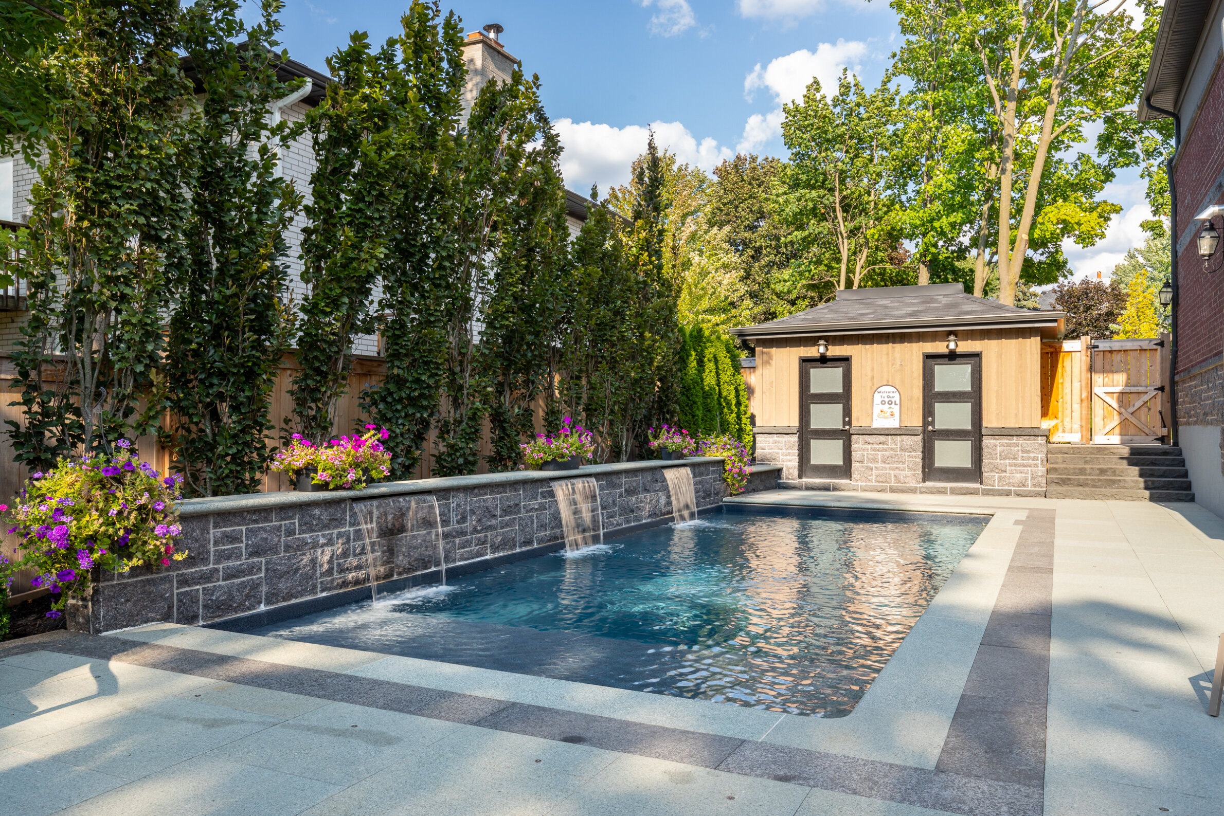 This is a luxurious backyard with a rectangular swimming pool, water features, a stone patio, flowering plants, a garden shed, and tall privacy trees.