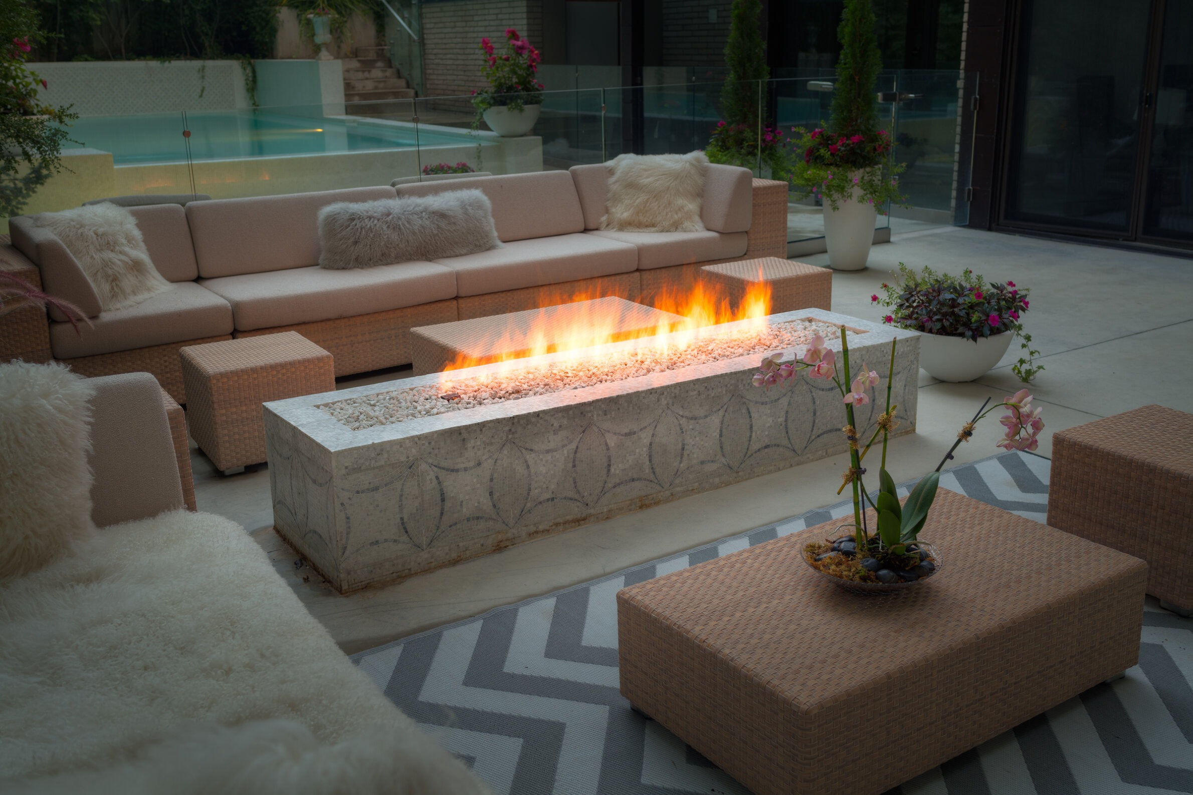 An outdoor patio area with a gas fireplace, cozy seating, patterned rug, potted plants, and a clear view over a calm swimming pool.