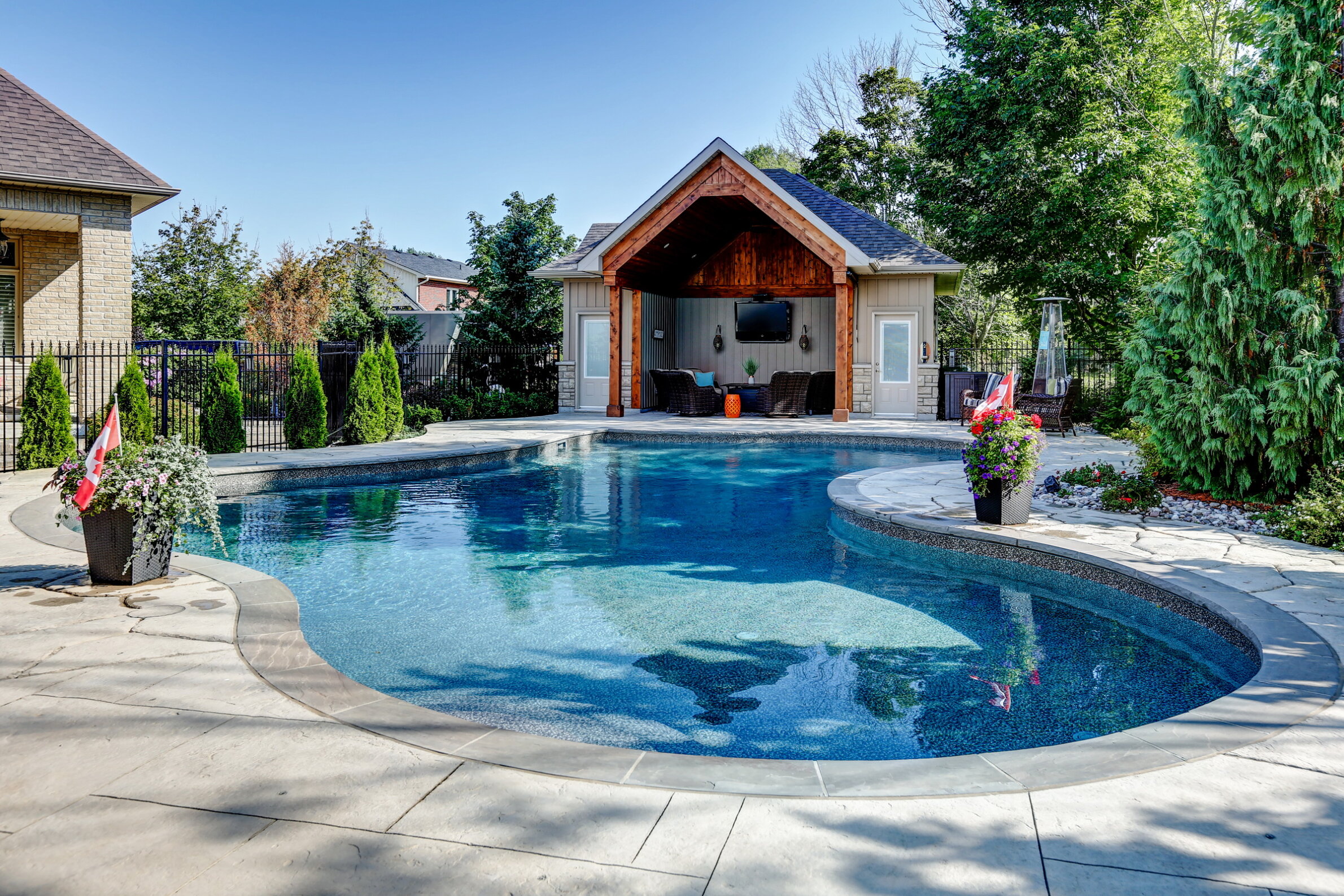A luxurious backyard with a curved swimming pool, a pool house, vibrant flowers, a Canadian flag, and green landscaping under a clear blue sky.