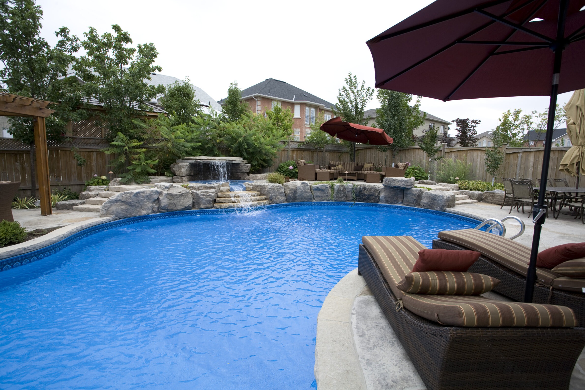 This image shows an inviting backyard with a curved swimming pool, waterfall feature, sun loungers, patio umbrellas, and landscaping against a wooden fence.