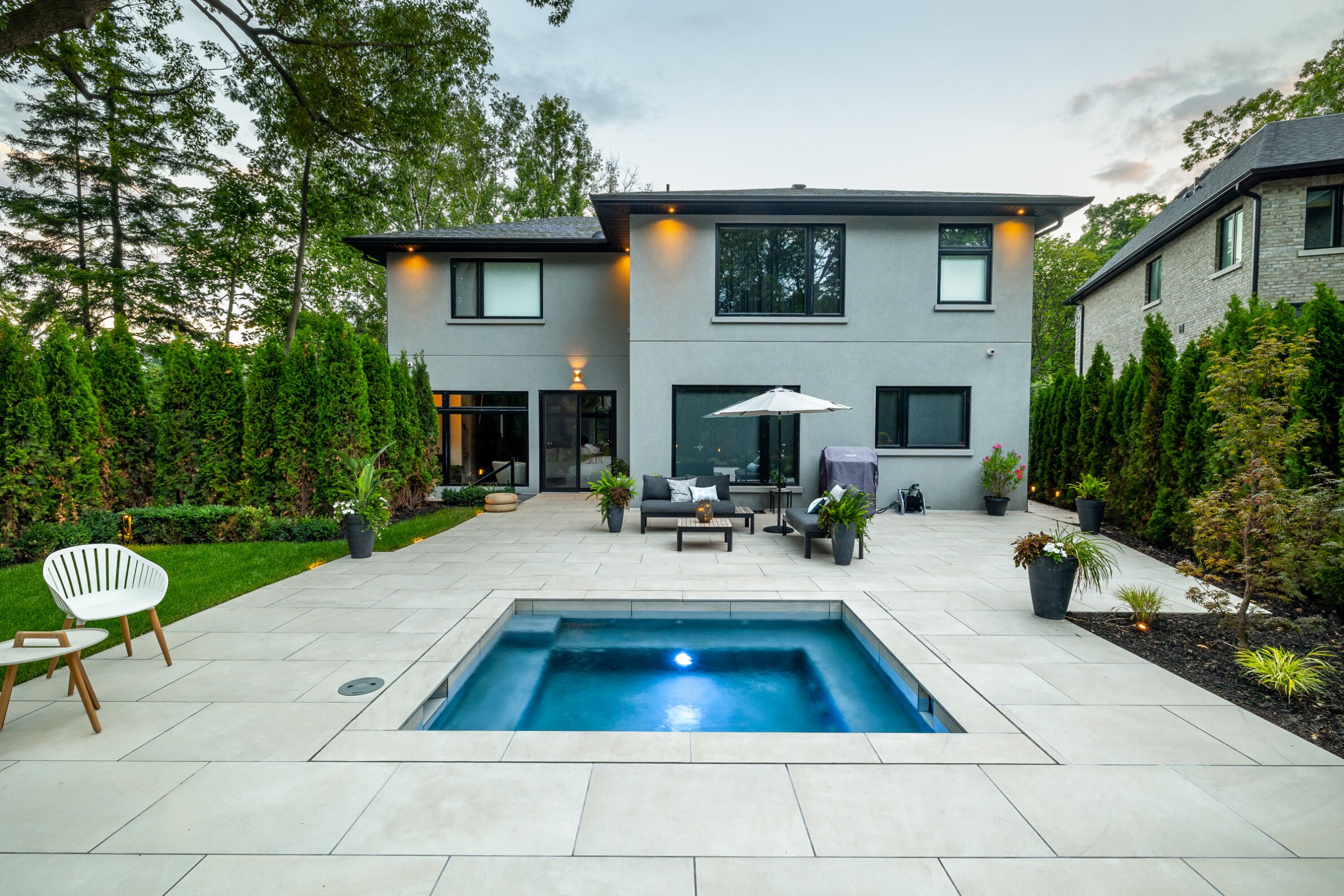 Modern two-story house with a flat roof, an outdoor seating area, and a small pool. Surrounded by lush greenery and a tiled patio space.