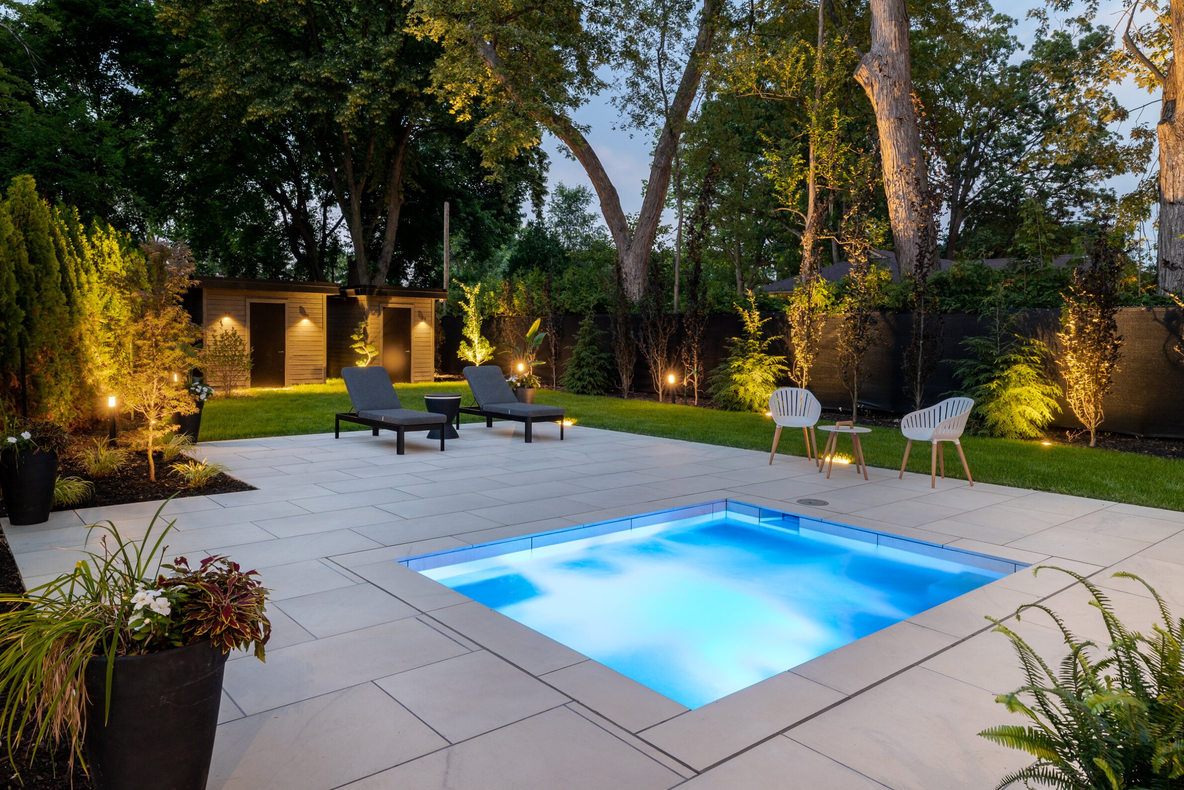 A luxurious backyard at dusk featuring a small illuminated pool, lounge chairs, elegant landscaping, lighting, and a modern shed surrounded by trees.